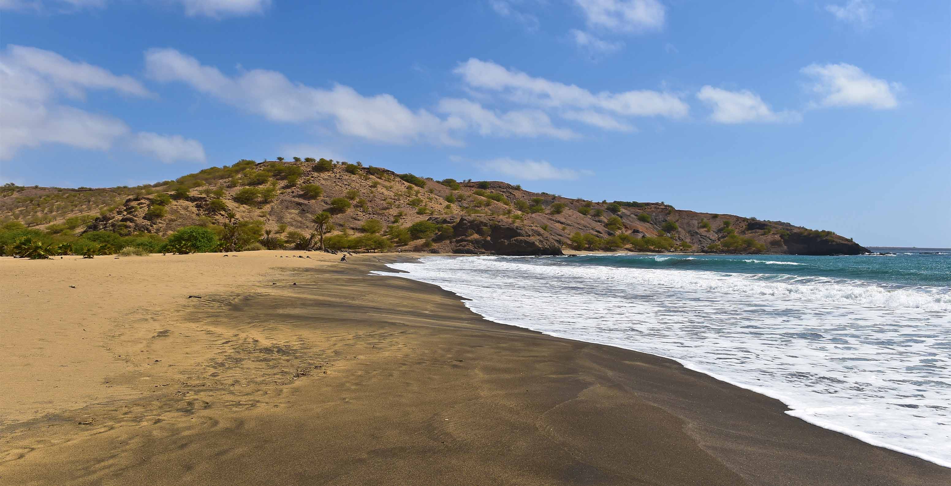 Strand mit dunklem Sand und klarem blauen Meer mit einigen Wellen an einem Tag mit wenig Wolken am Himmel