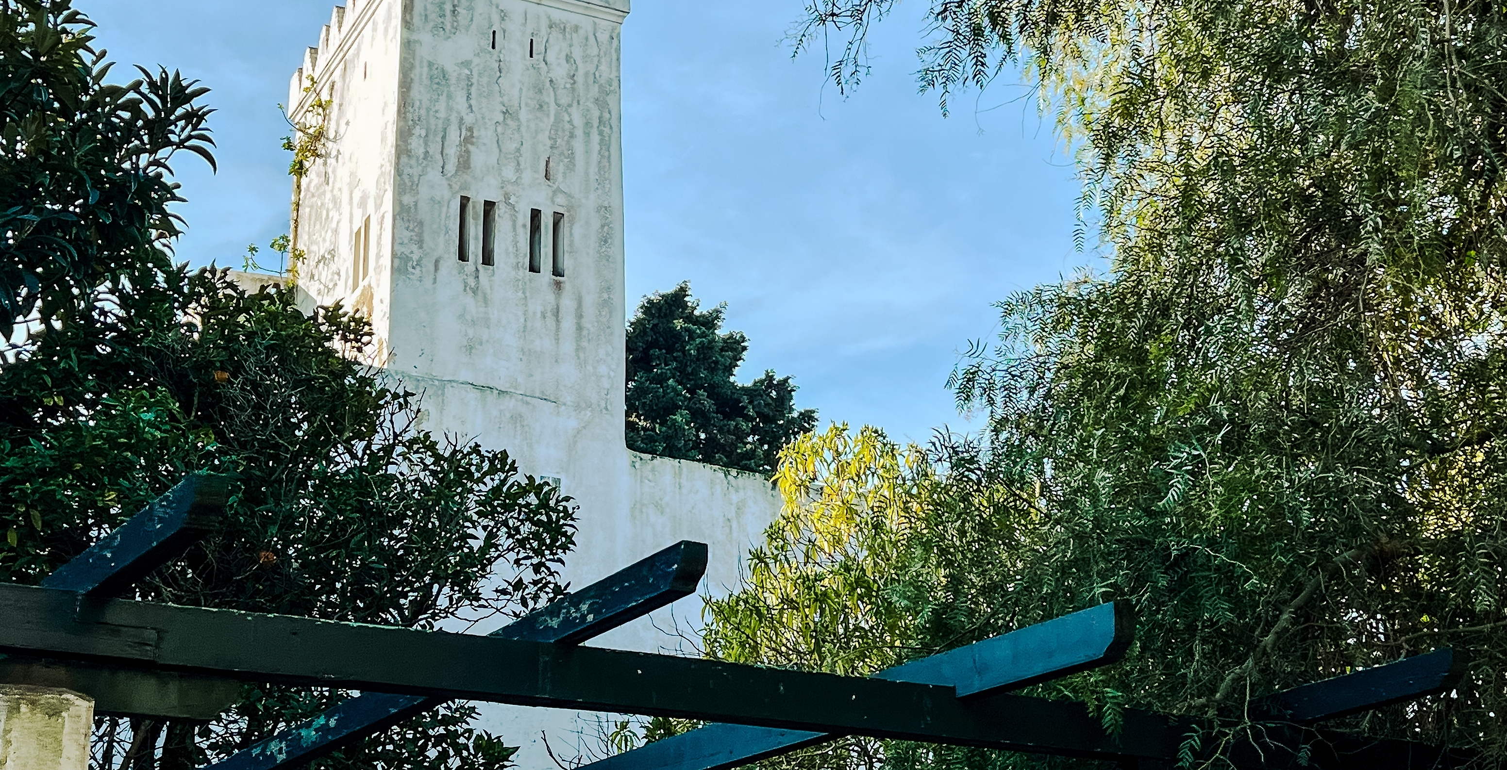 Blick auf einen weißen Turm mit typischen marokkanischen Merkmalen, versteckt zwischen Bäumen, unter einem blauen Himmel