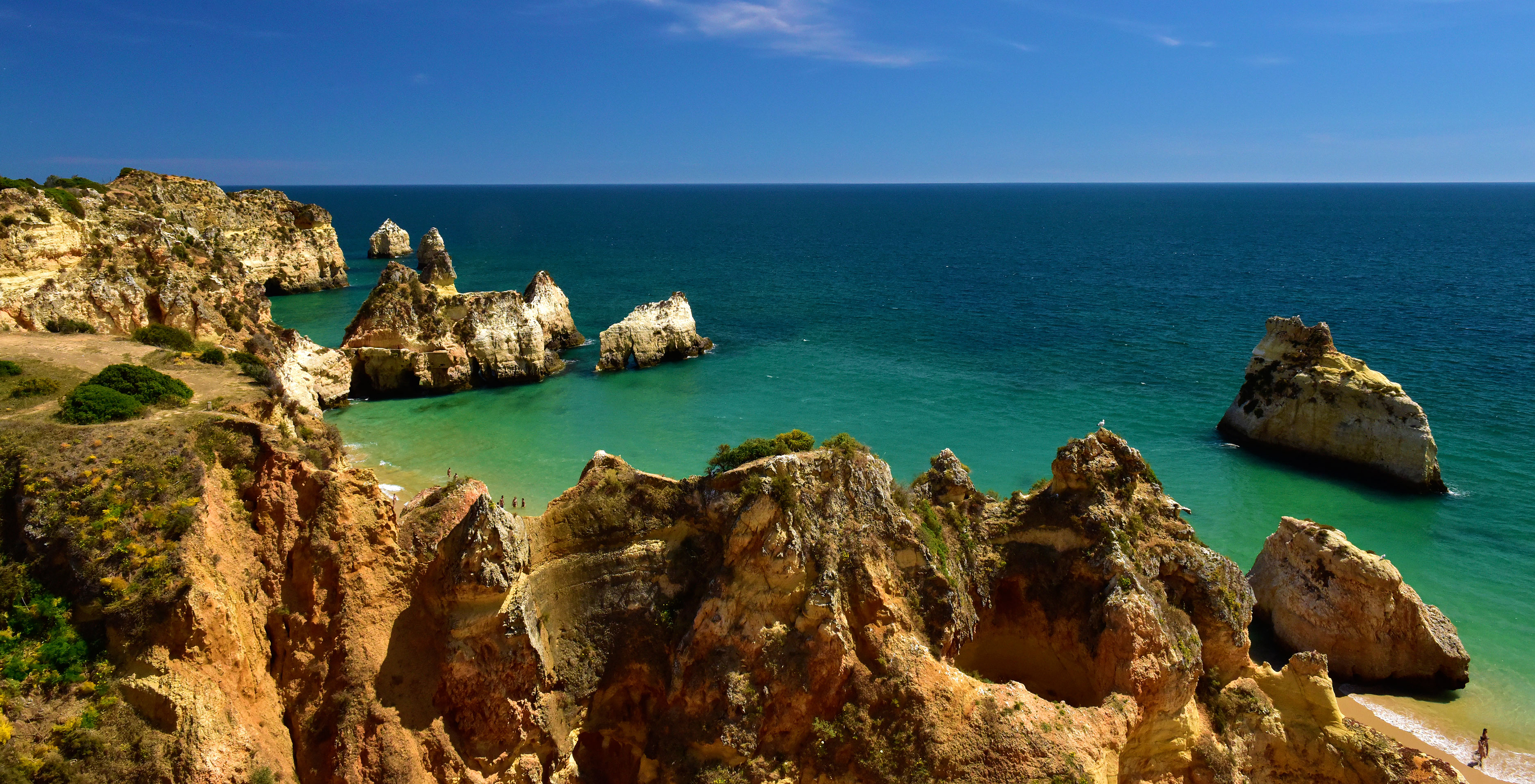 Blick auf die Klippe zu einem Strand in Alvor mit klarem blauem Wasser im Hintergrund und blauem Himmel mit wenigen Wolken