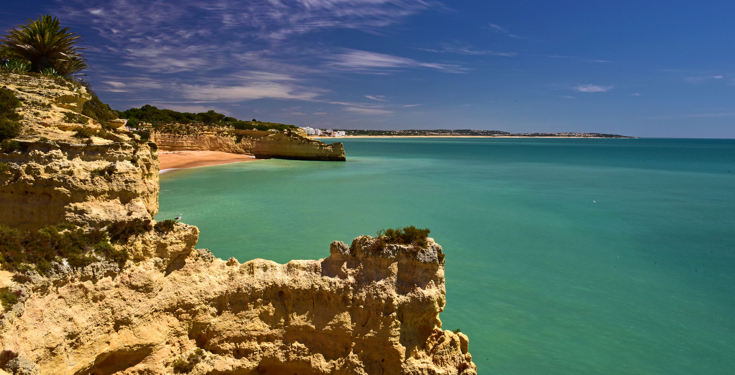 Blick von der Klippe auf einen Strand im Alvor mit klarem blauen Wasser im Hintergrund und blauem Himmel mit wenigen Wolken