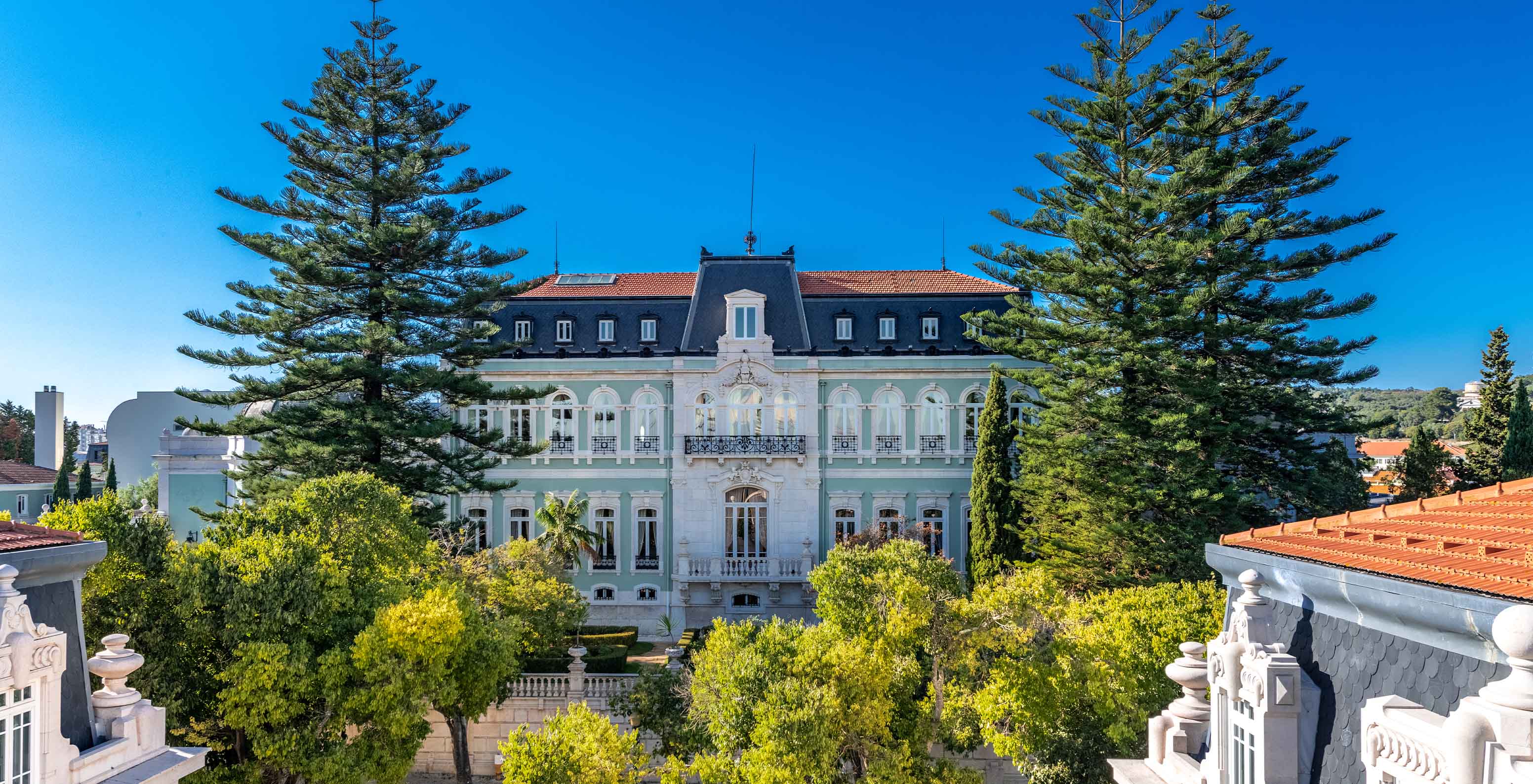 Blick auf die Vorderseite des Hauptgebäudes des Pestana Palace Lisboa, mit zwei großen Bäumen, die das Hotel umrahmen