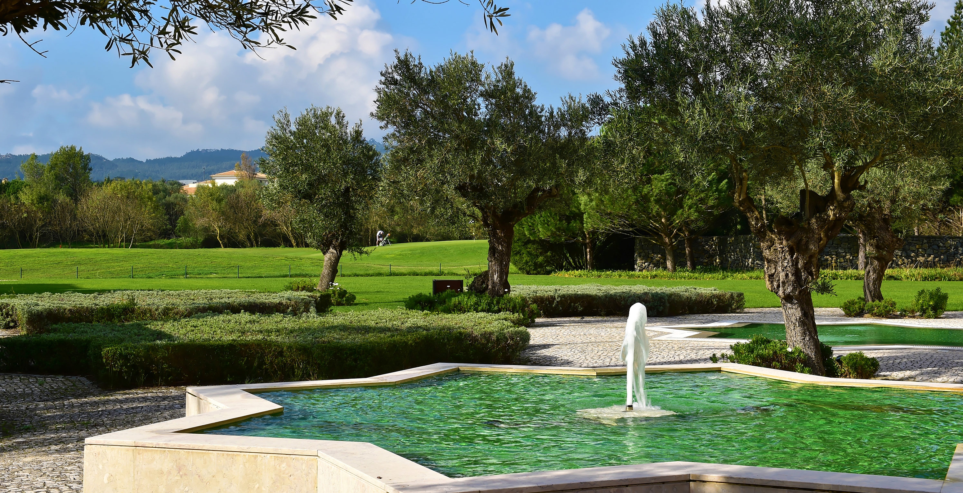 Brunnen in einem sternförmigen Teich im Pestana Sintra Golf, mit Garten und Bäumen im Hintergrund an einem sonnigen Tag