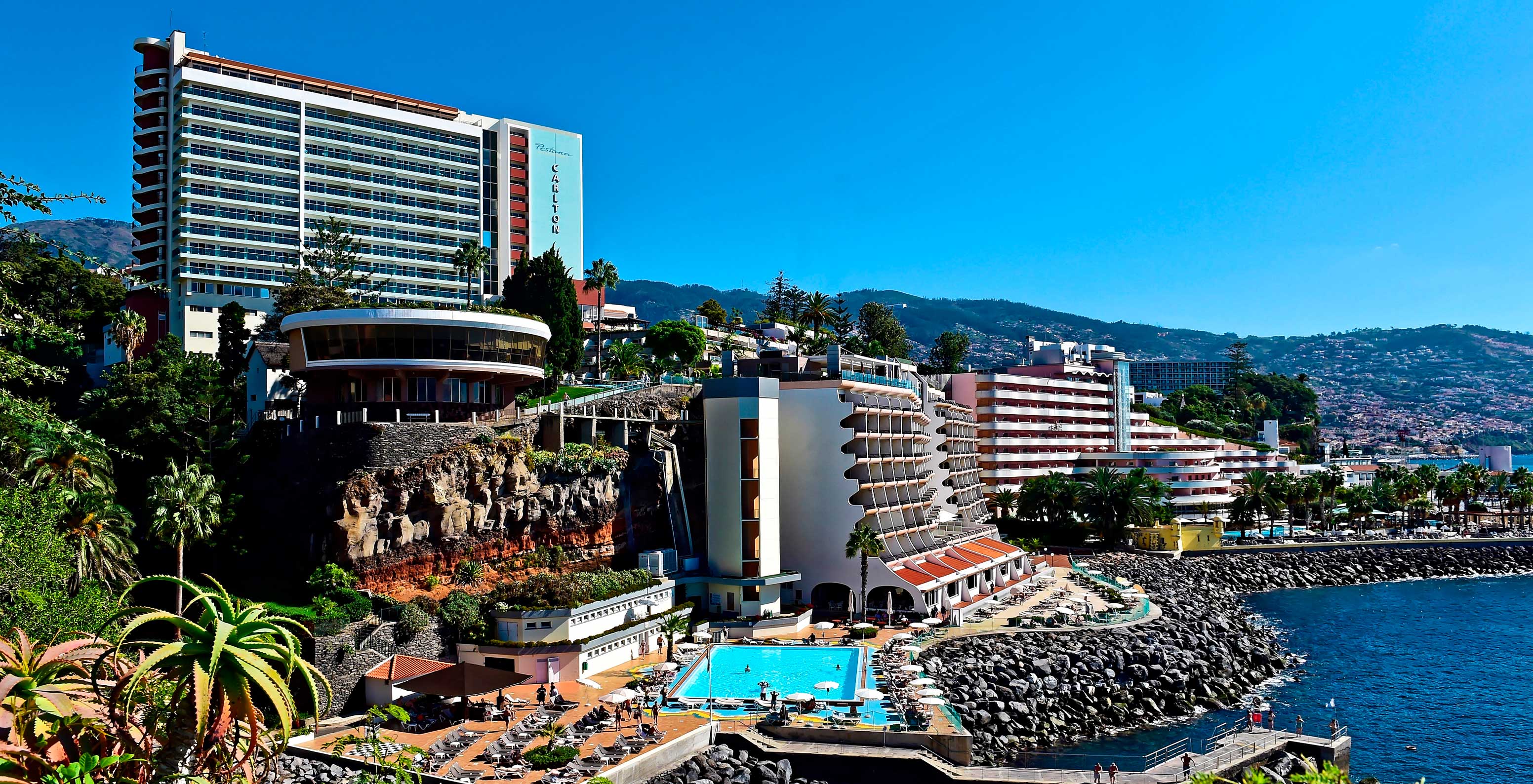 Blick vom Meer auf das Pestana Carlton Madeira, mit großen Gebäuden und Pool, umgeben von Natur