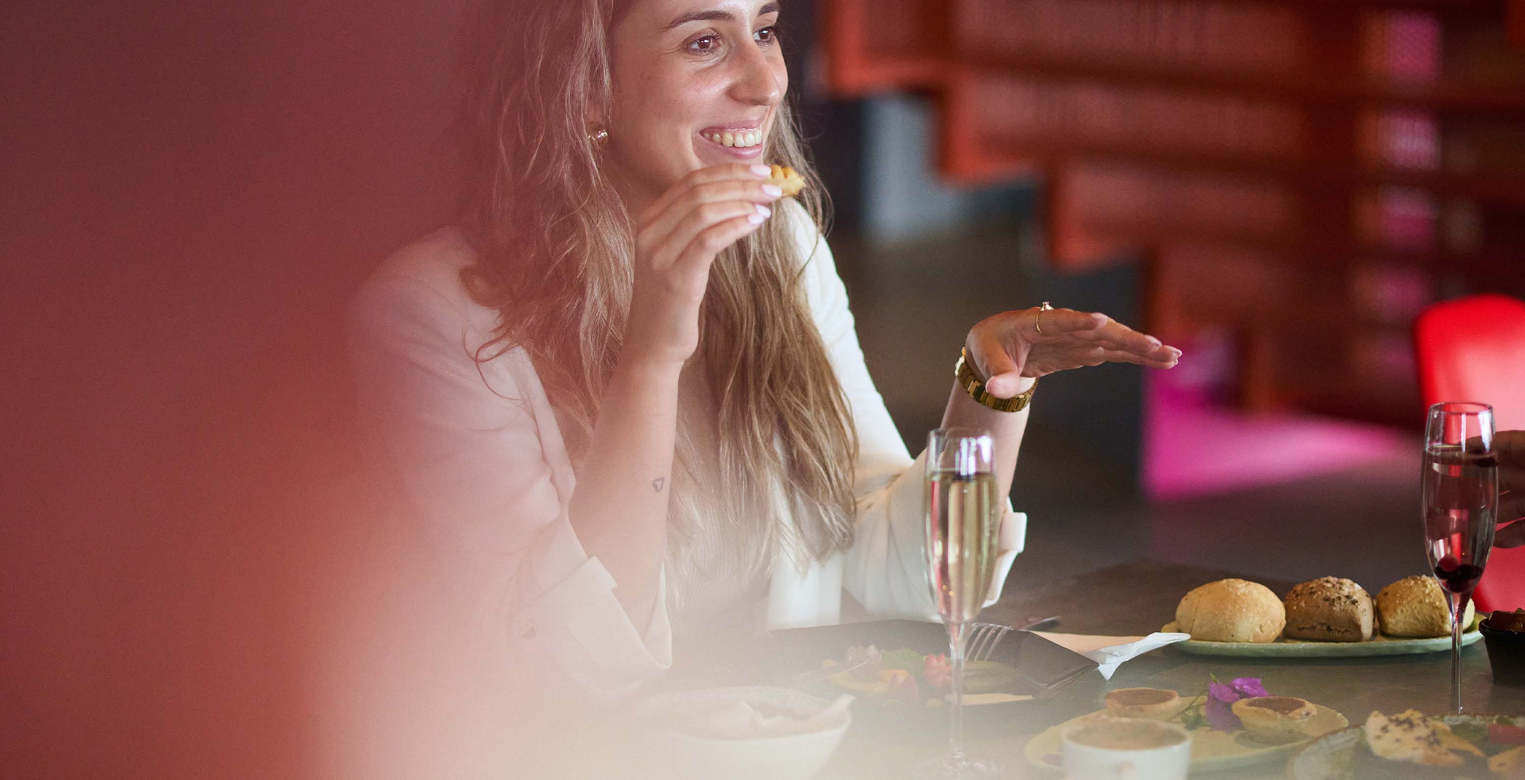 Lächelnde Frau isst einen Snack, sitzt an einem Tisch mit mehreren Kuchen und Snacks, begleitet von einem Glas Sekt