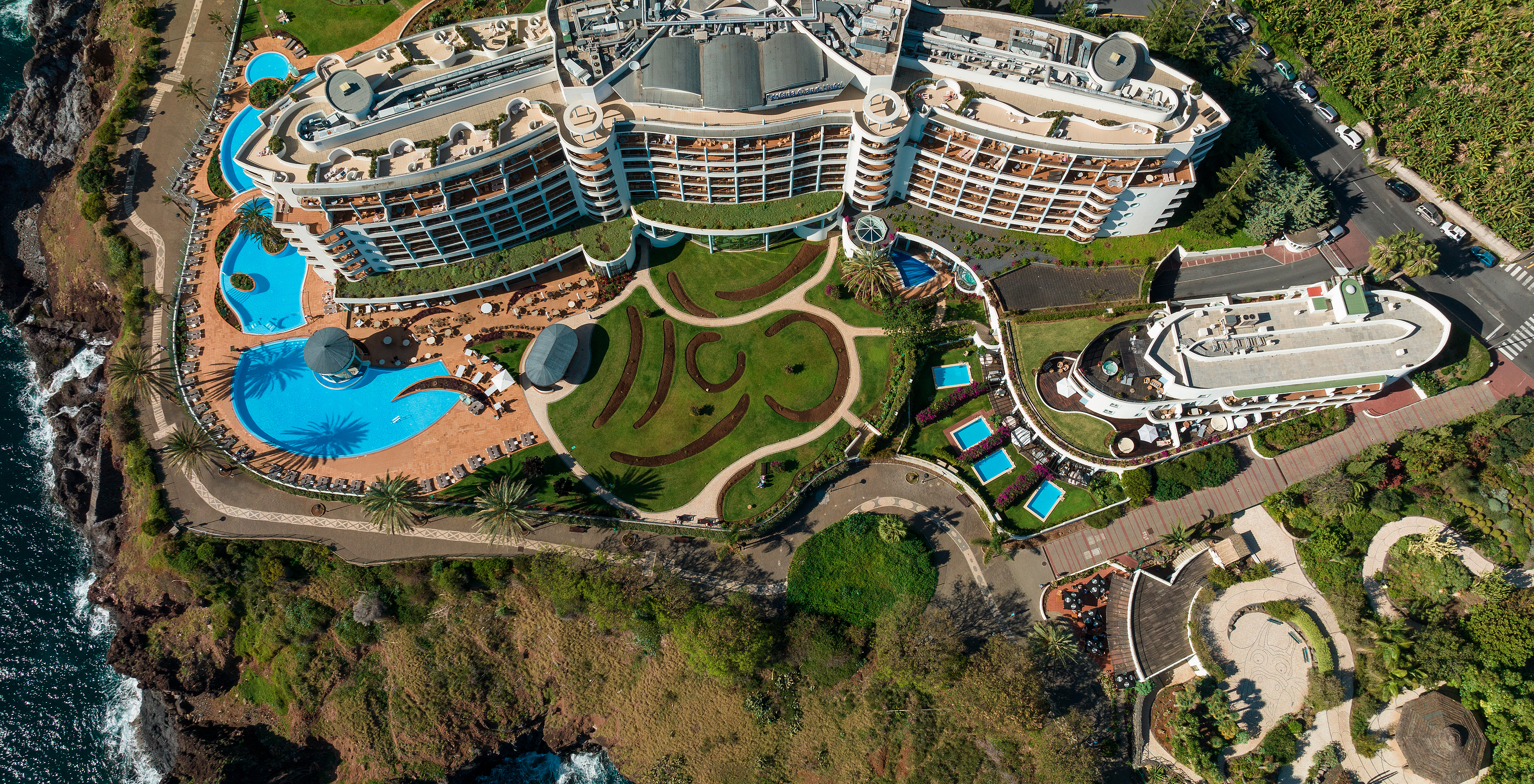 Blick von oben auf das Pestana Grand, mit Außenpool, Gärten und Fassade des 5-Sterne-Hotels am Meer