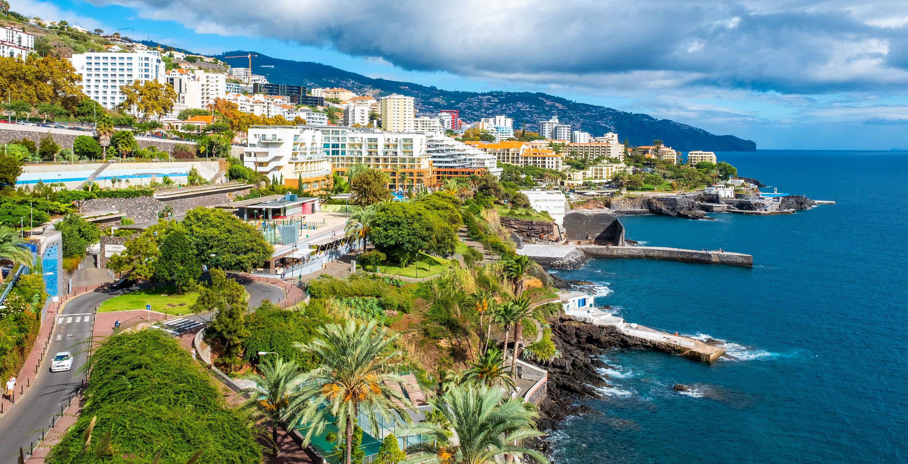 Blick auf die Stadt Funchal, am Meer mit mehreren Gebäuden, Vegetation und einer Straße mit einem vorbeifahrenden Auto