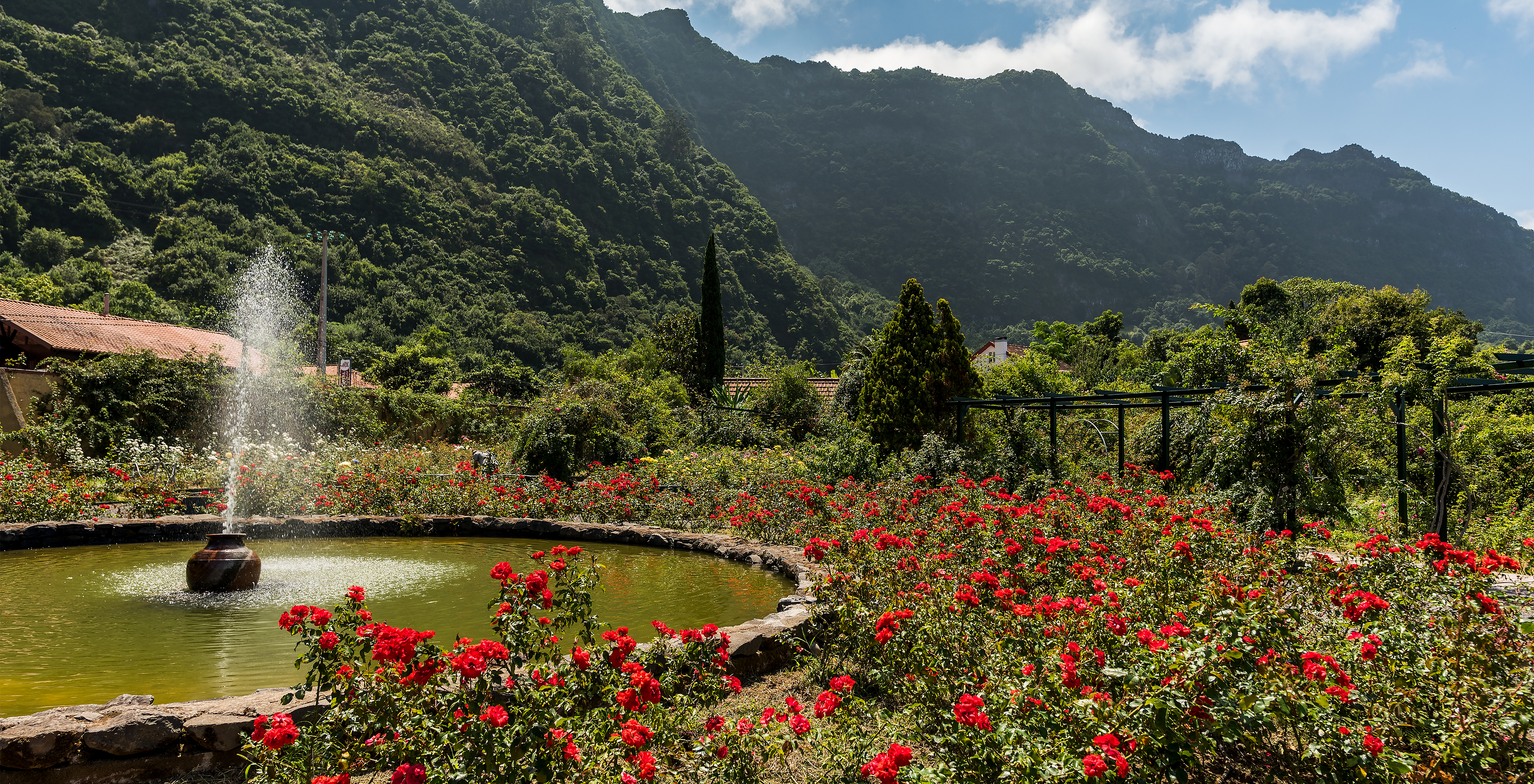 Brunnen im Zentrum des Rosengartens der Quinta do Arco, mit viel Vegetation und roten Rosen, im Hintergrund die Berge