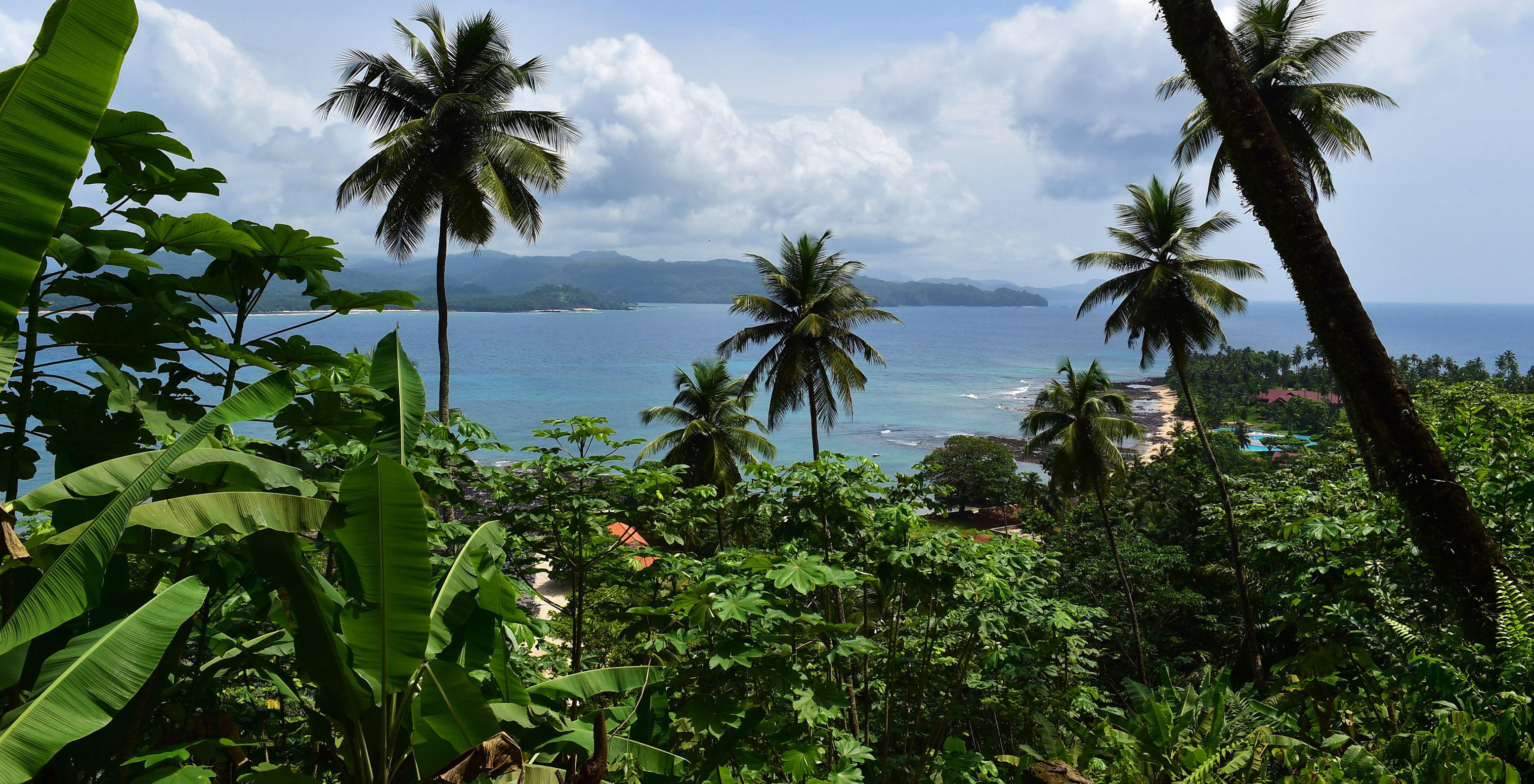 Blick auf den Pestana Equador Ilheu das Rolas mit tropischer Vegetation wie Palmen, blauem Meer und Bergen im Hintergrund