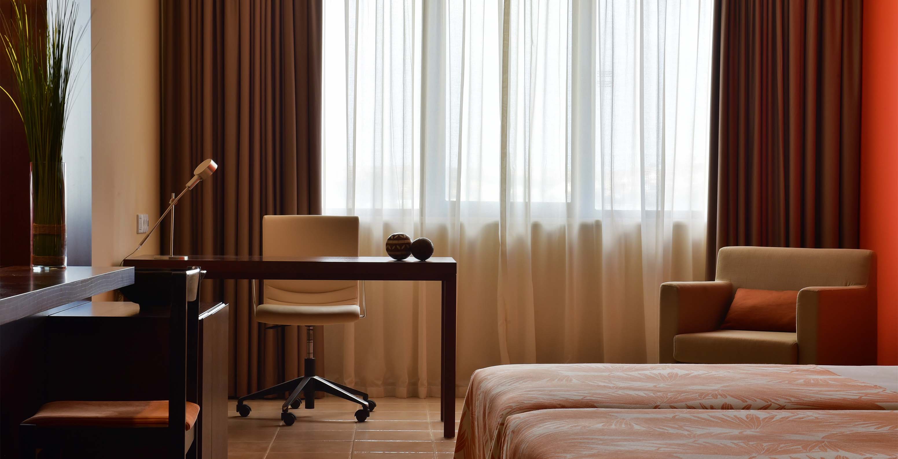 The Standard room at Pestana Tropico features a desk with lamp and a chair, and an armchair