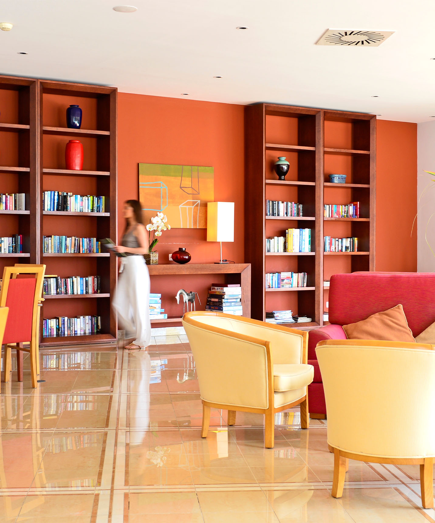 Hotel lounge with sofas, chairs, and armchairs, bookshelves and decor, with a lady walking in the back