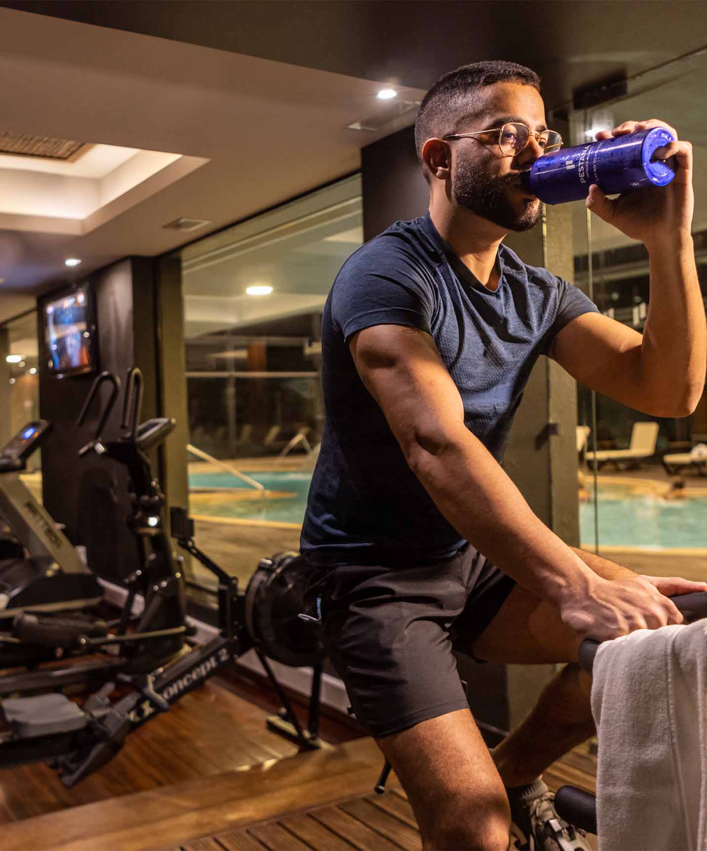 Gym with a man drinking water on a bike, with ellipticals and treadmills, wood floor next to indoor pool