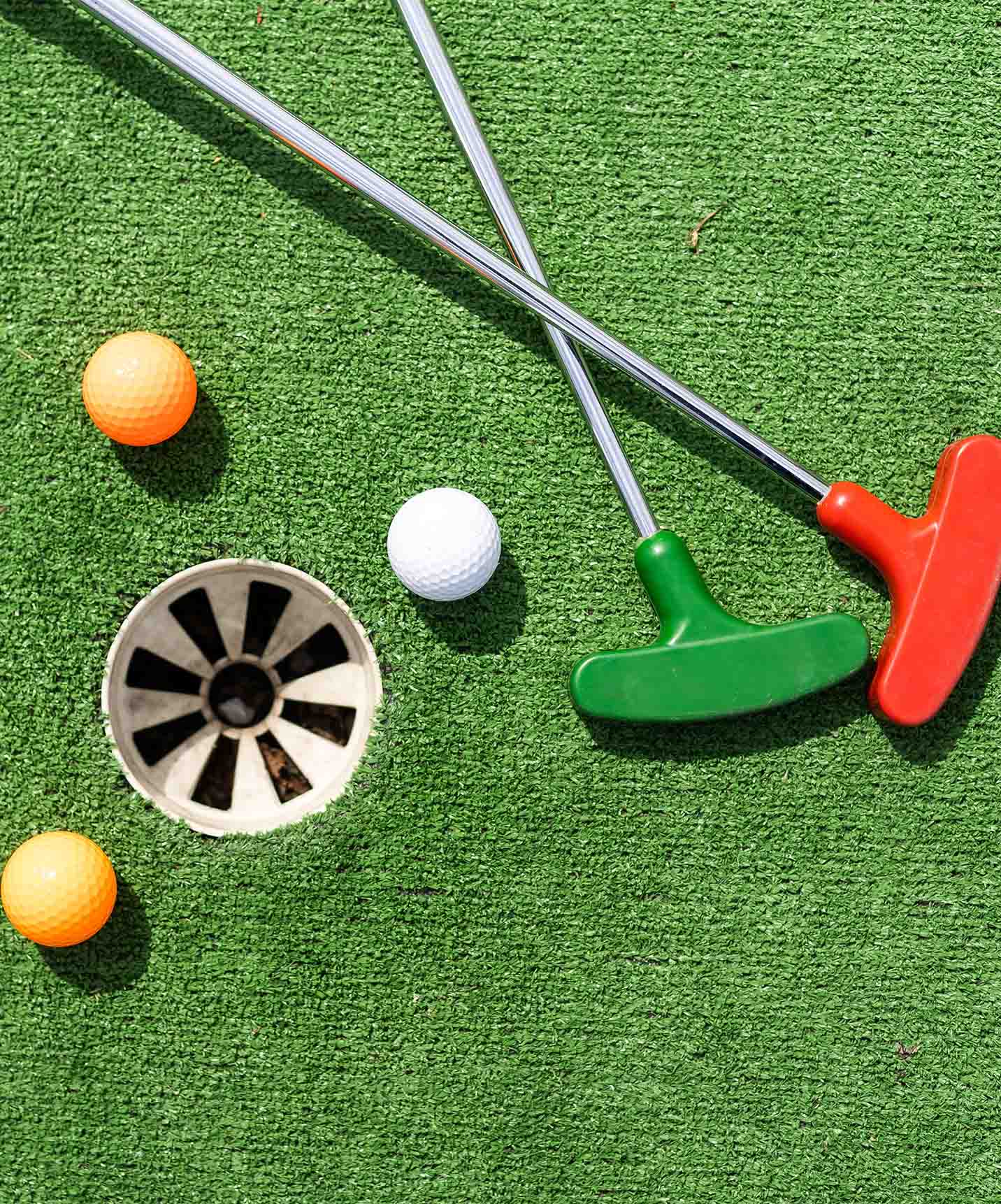 Mini-golf hole with green grass background, red and green clubs, two orange golf balls and one white