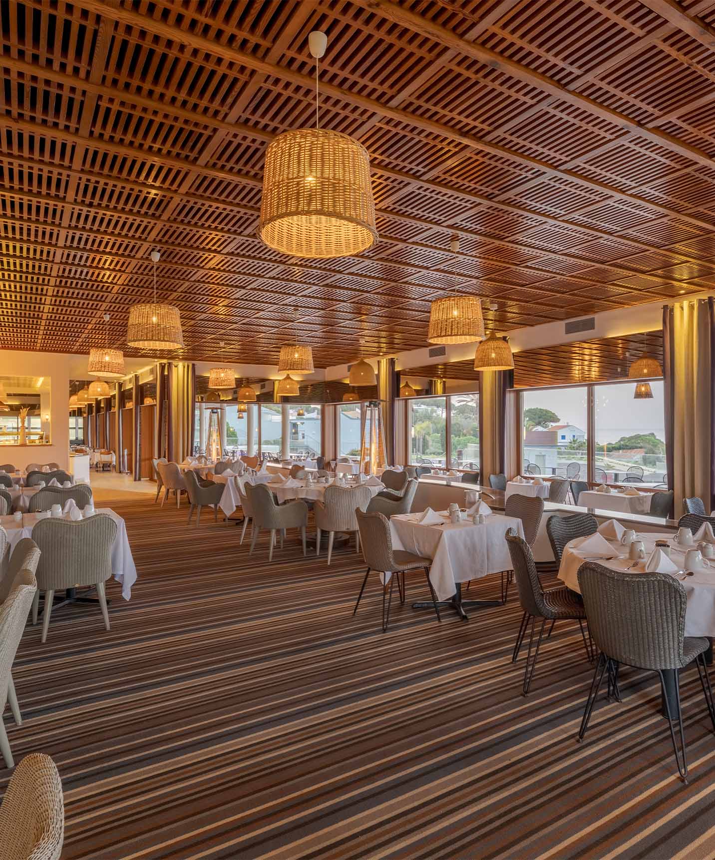 Restaurant with square and round tables with ceiling lamps, striped carpet and wooden ceiling, sea view