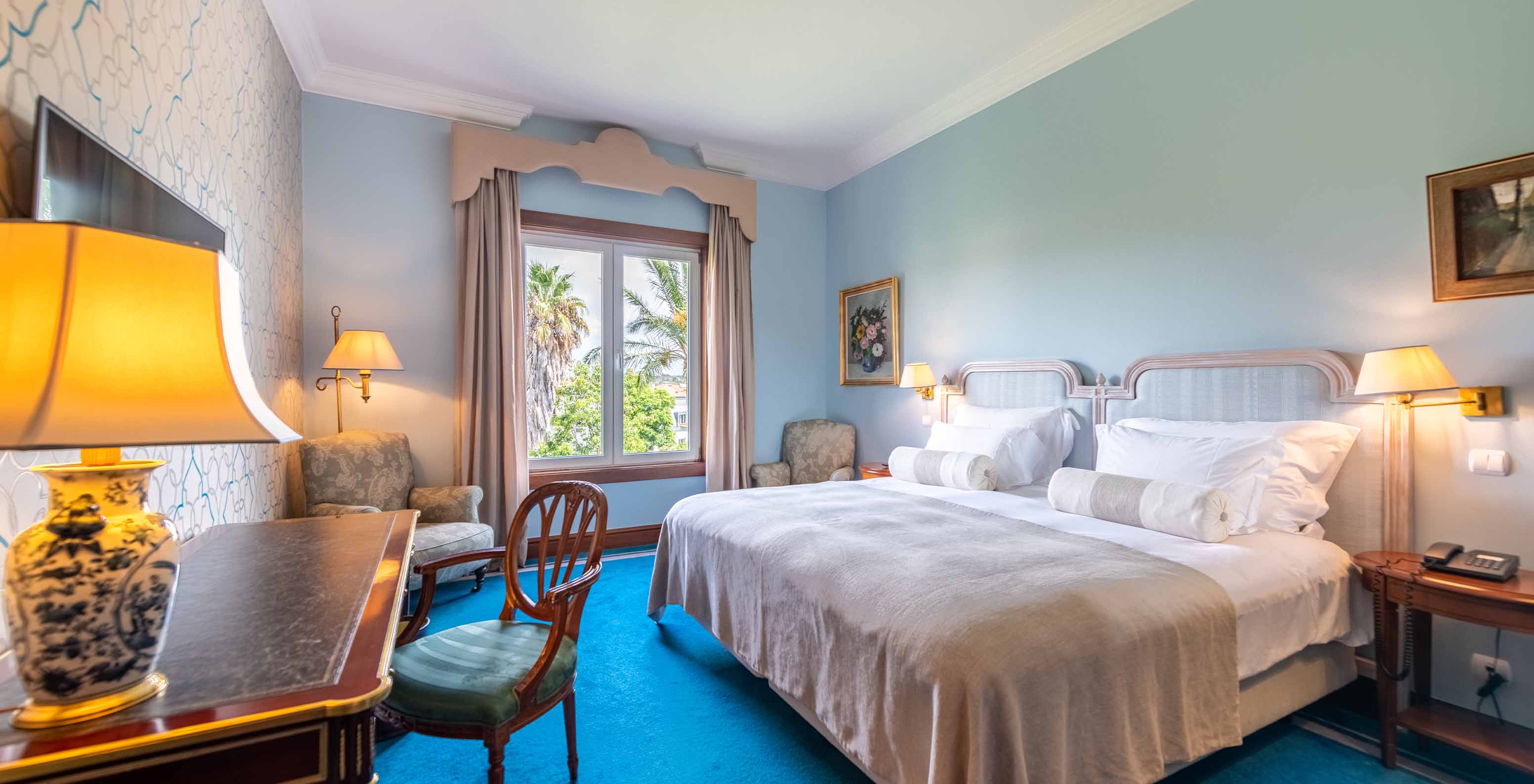 The Deluxe Garden room at Pestana Palace Lisbon has a double bed, two bedside tables, a desk, and a window with a garden view