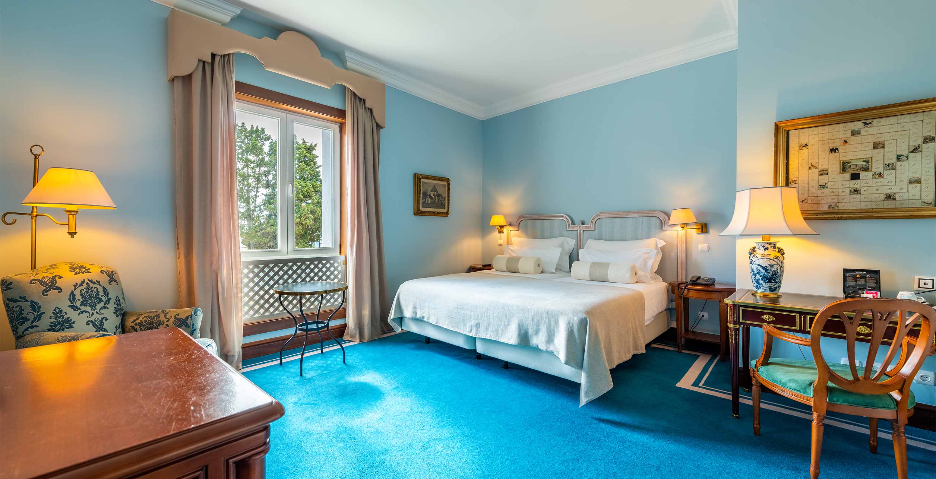 The Deluxe room at Pestana Palace Lisbon has two single beds, a window, an armchair, a desk, and a blue carpet