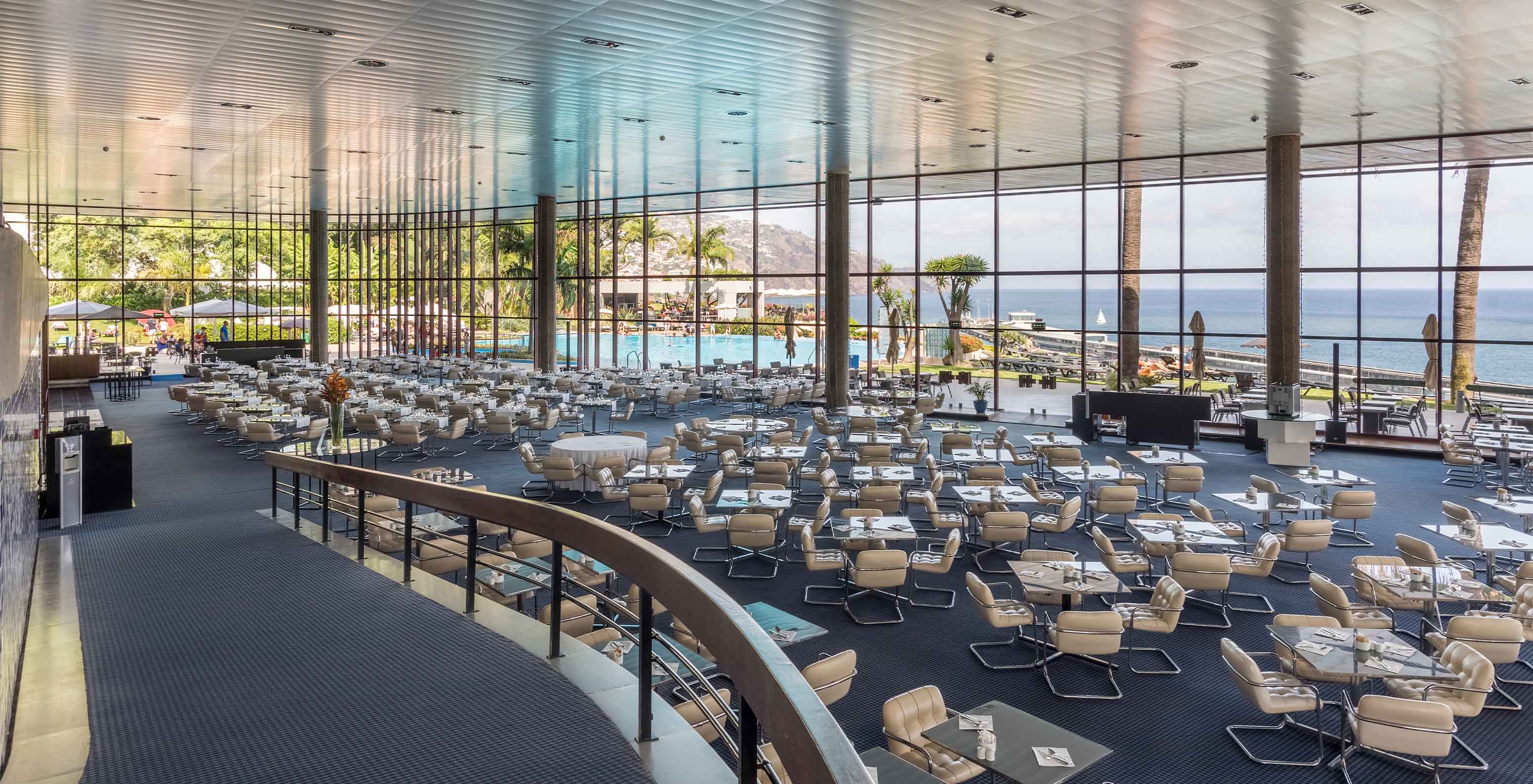 Panoramic view of a restaurant with several tables, a ramp, and a large window overlooking the pool and sea