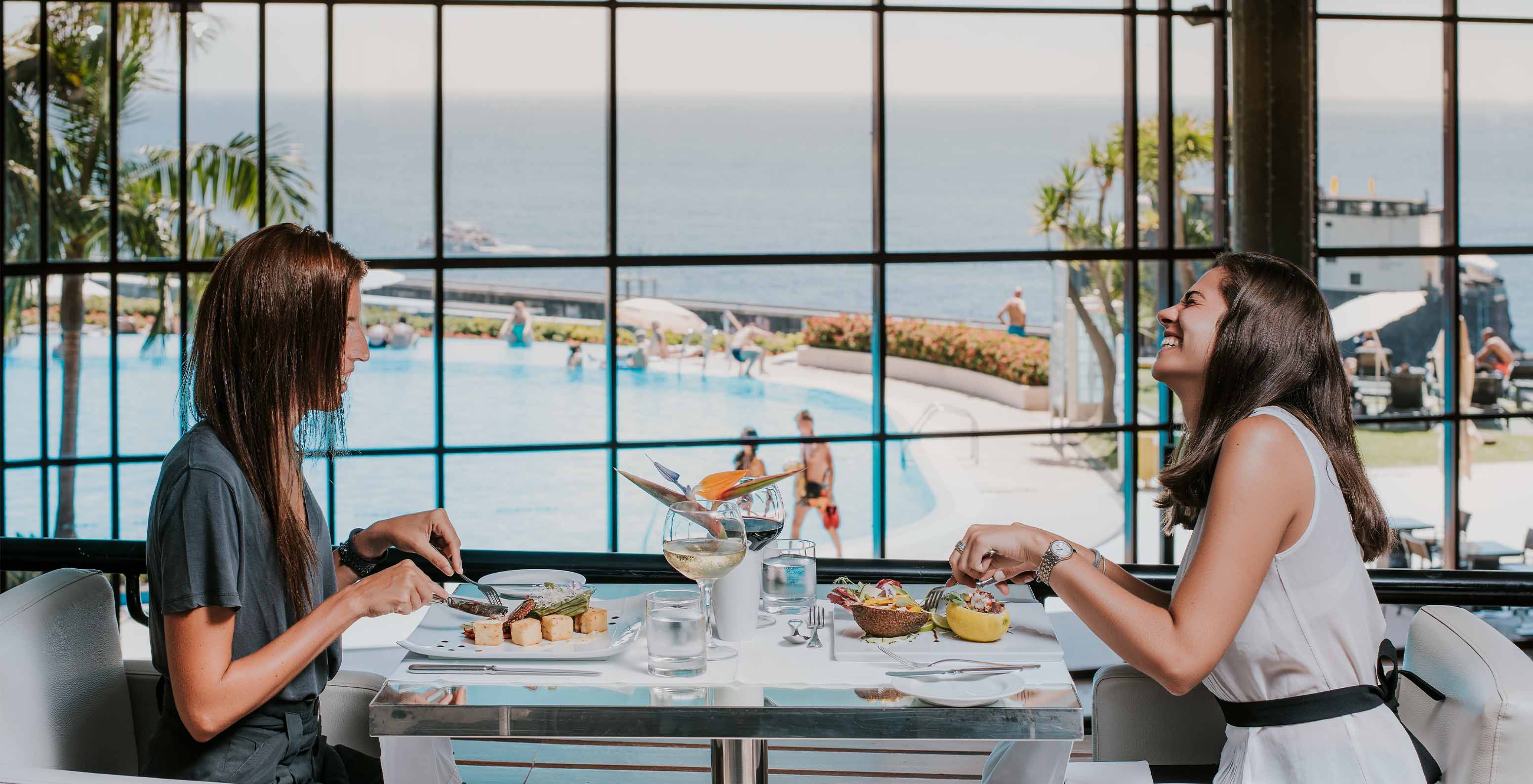 Two friends having lunch indoors at Pestana Casino Park, with a view of the pool and the sea