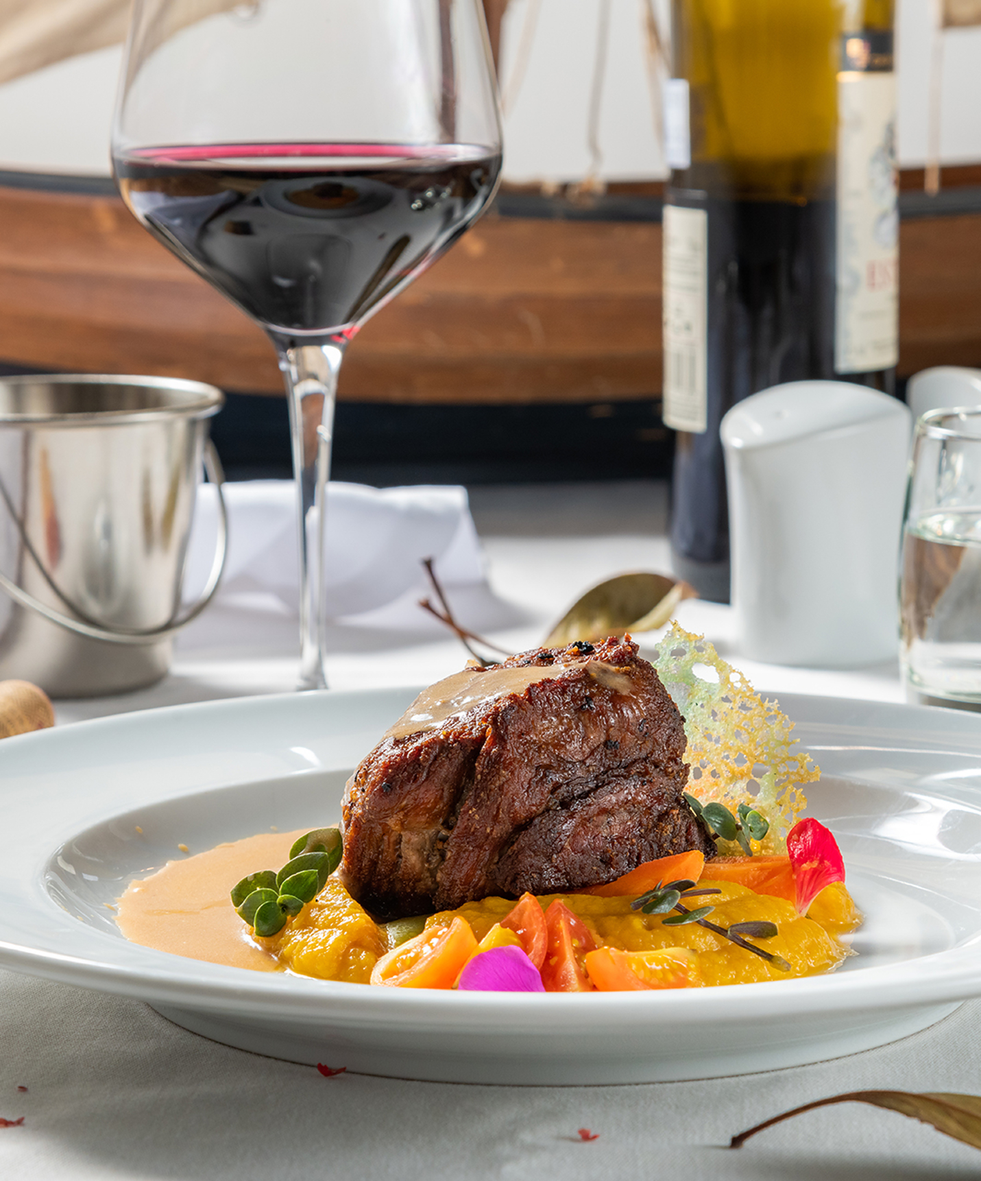 Meat dish on a bed of puree, with cherry tomatoes and decorative elements, accompanied by a glass of wine and water