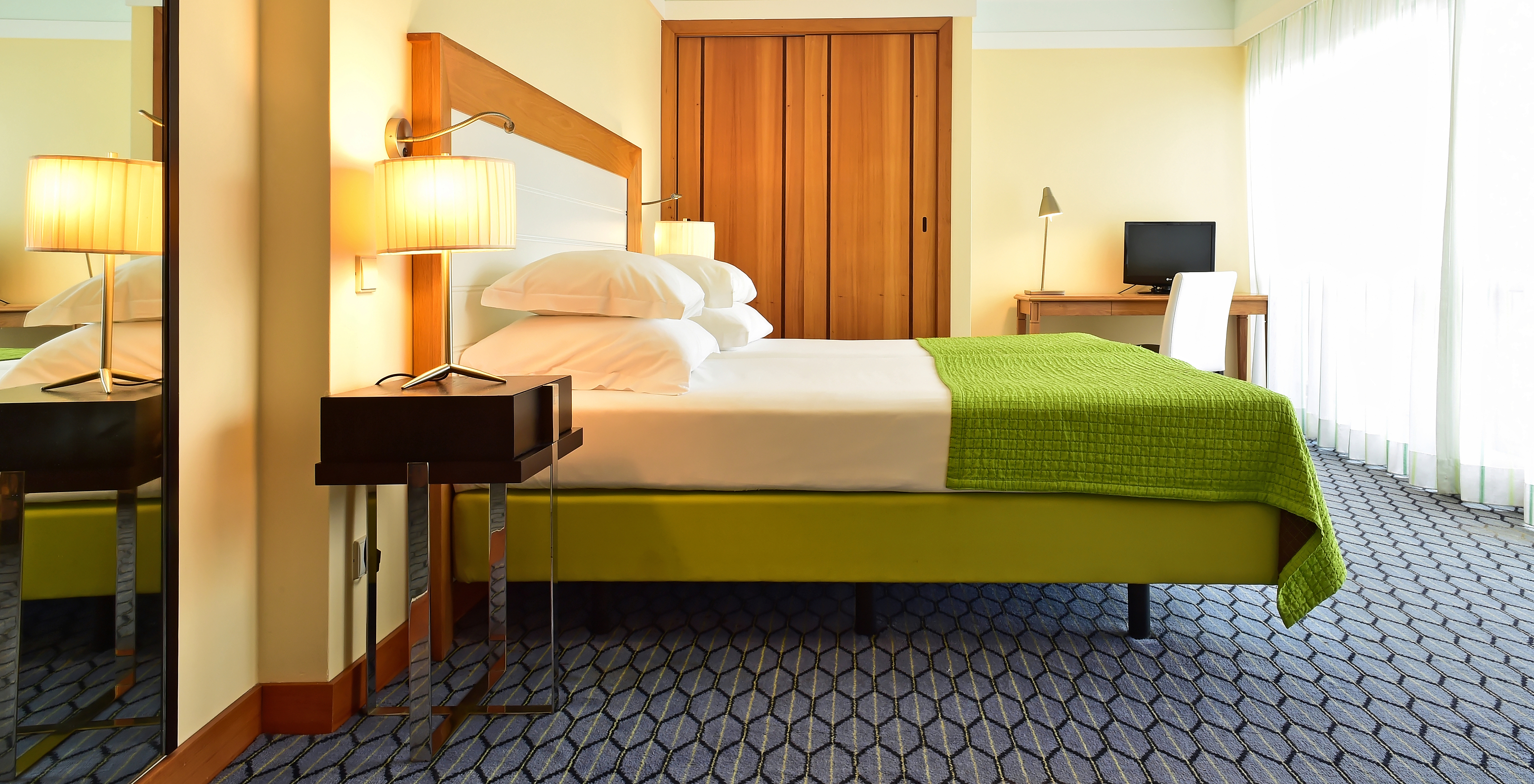 The Junior Suite of Pestana Promenade has a double bed with a green bedspread, a wardrobe, and a desk