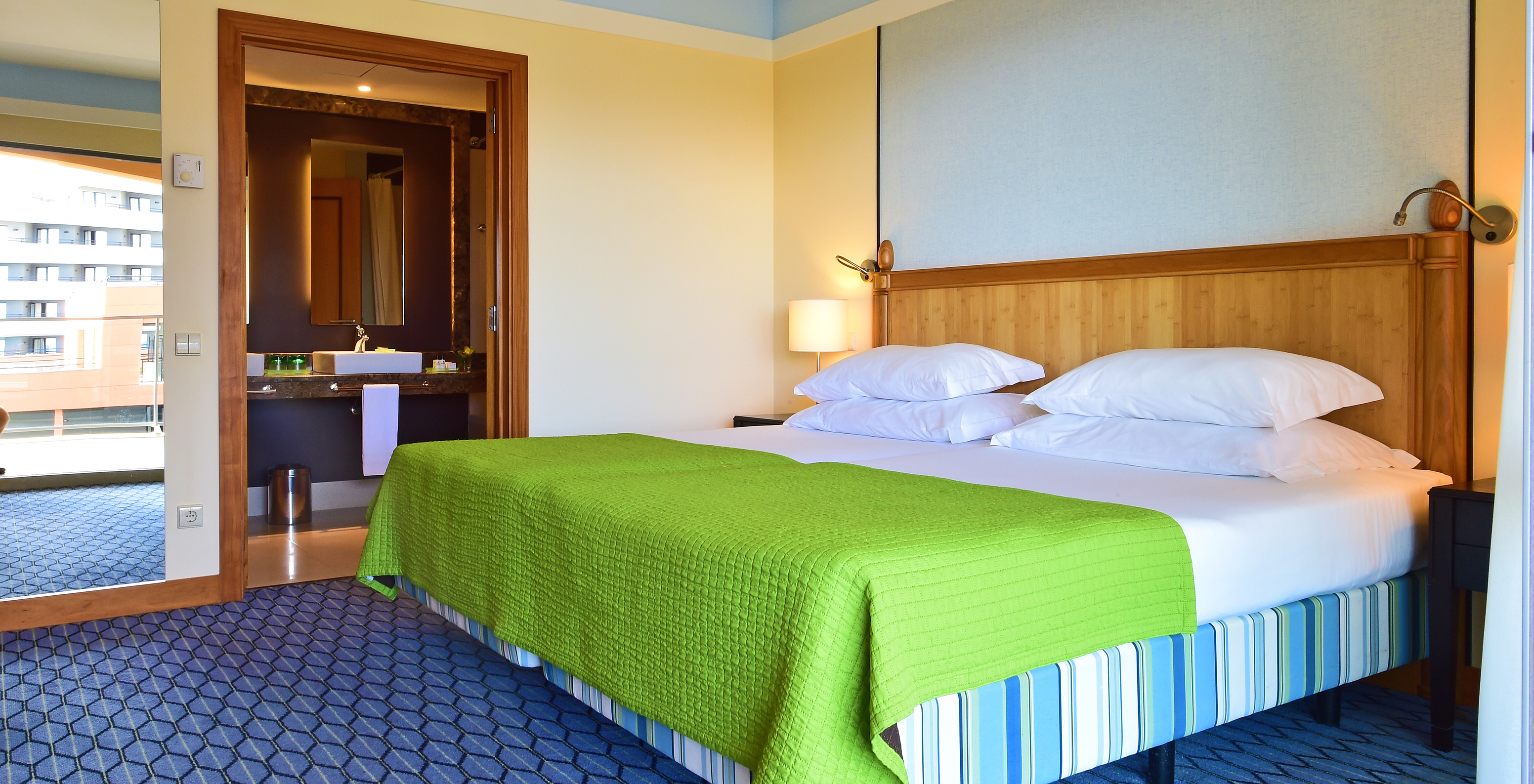 The Suite of Pestana Promenade has a double bed with a green bedspread, a tall mirror, and a bathroom