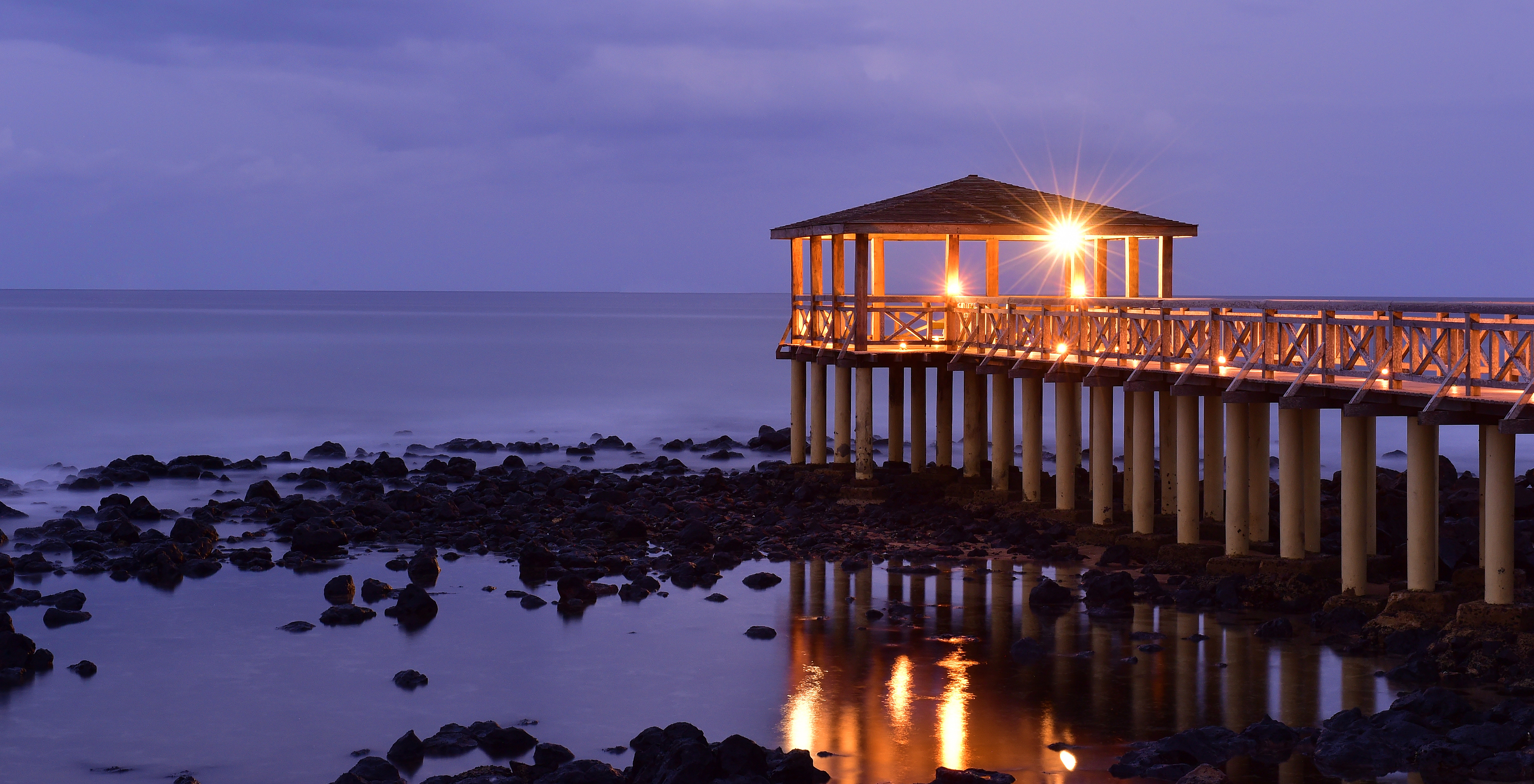 Side view of the wooden pier over the sea at sunset, illuminated by the lights of the structure