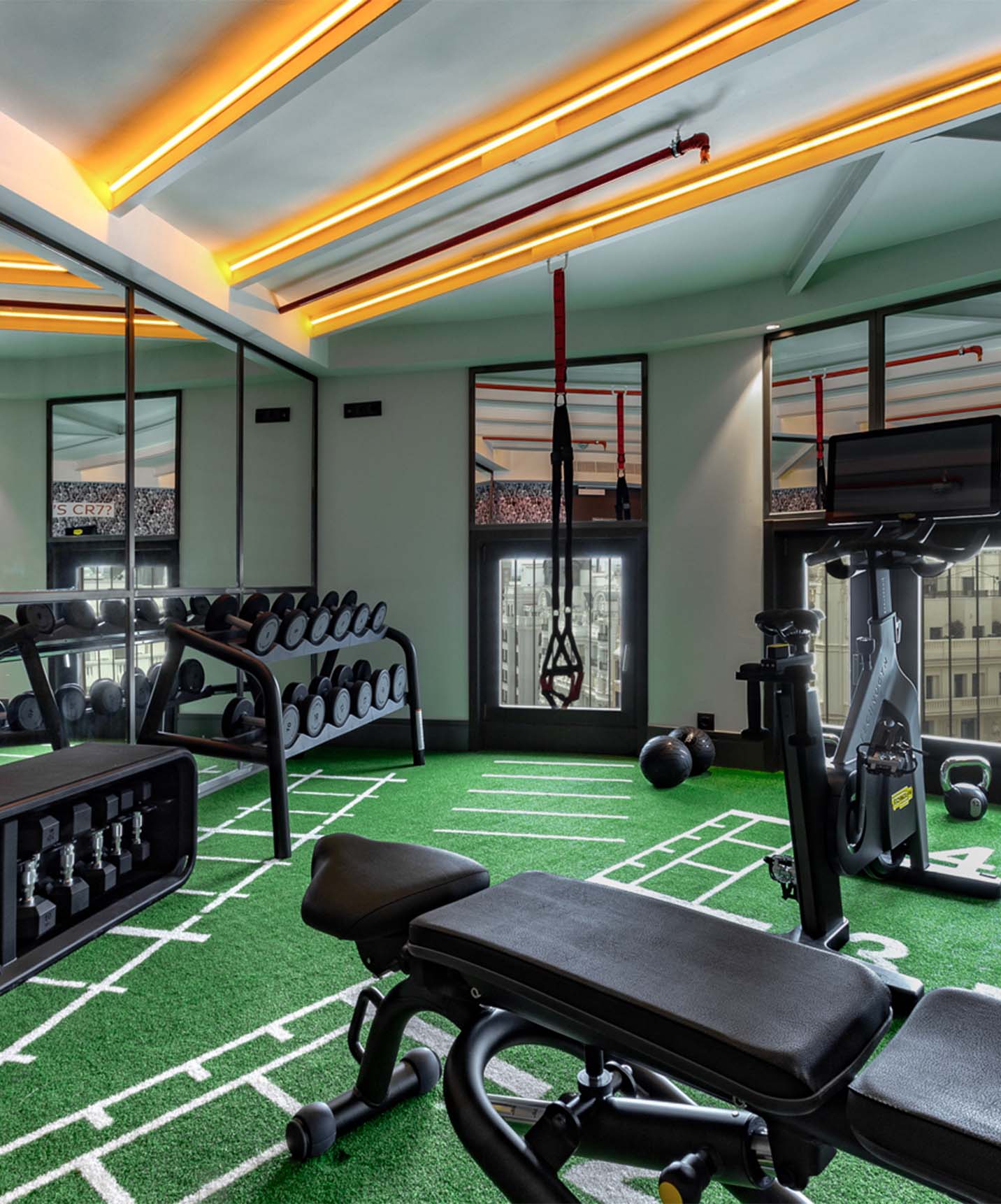 Gym with dumbbells, bike, weight bench, and TRX with mirrors and large windows overlooking front buildings