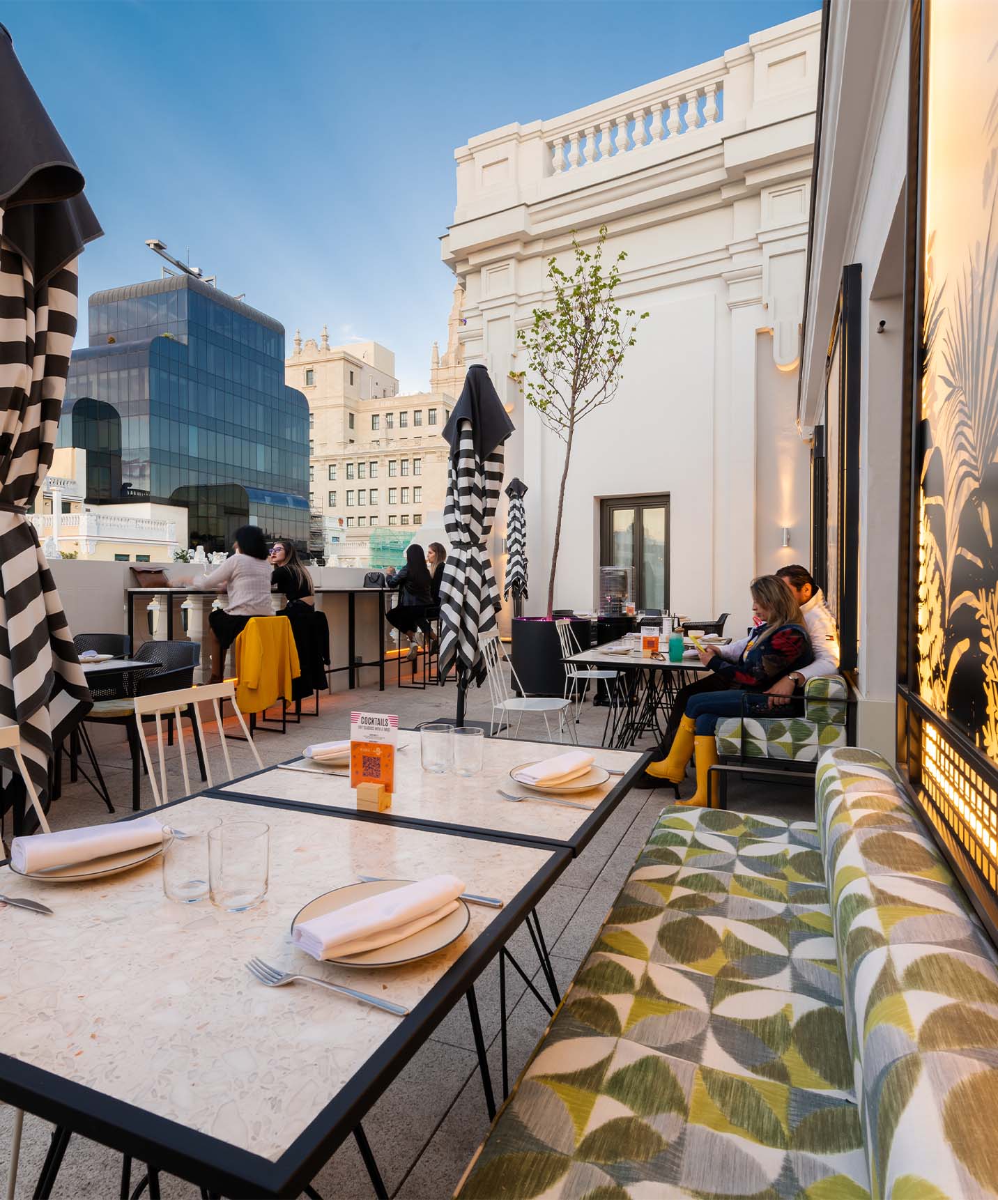 Outdoor part of the restaurant with sofas, chairs, tables, and umbrellas on the rooftop with views of the front buildings