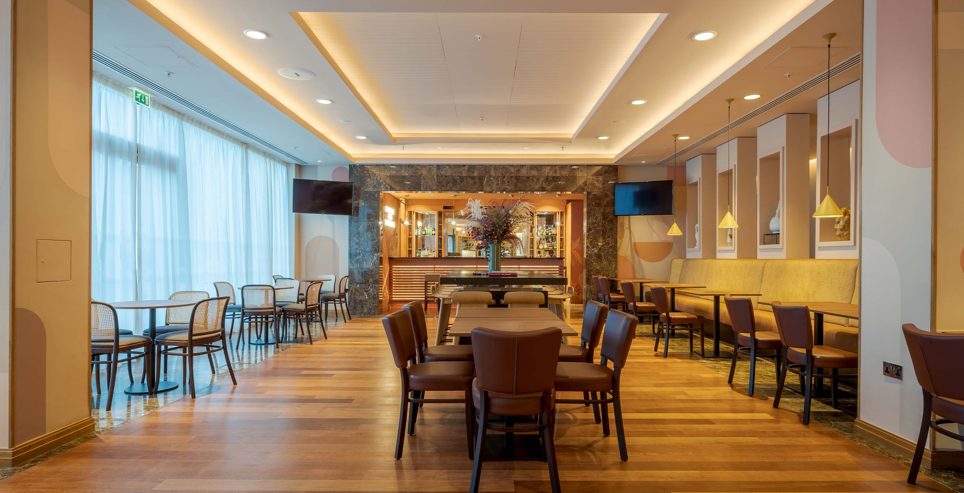 Hotel bar area with various tables, chairs, sofas, TVs, ceiling lamps, and the counter in the background