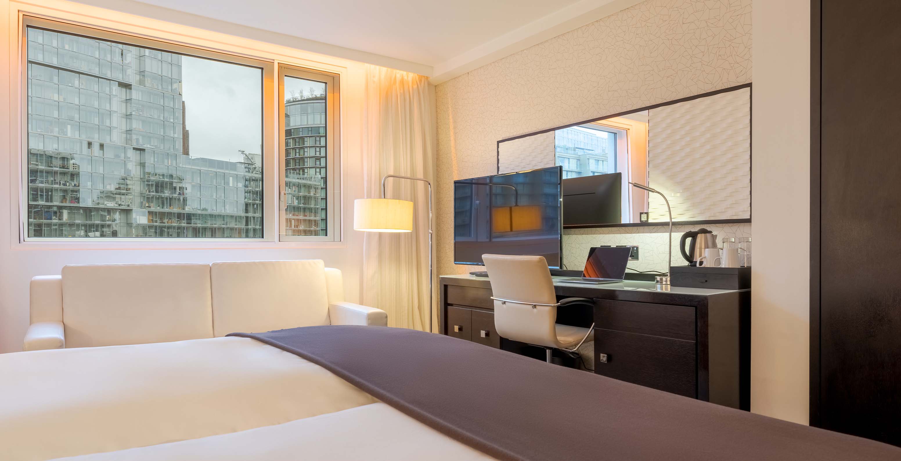 The Deluxe Family at Pestana Chelsea Bridge has two joined beds, a desk with a TV, a sofa, and city windows