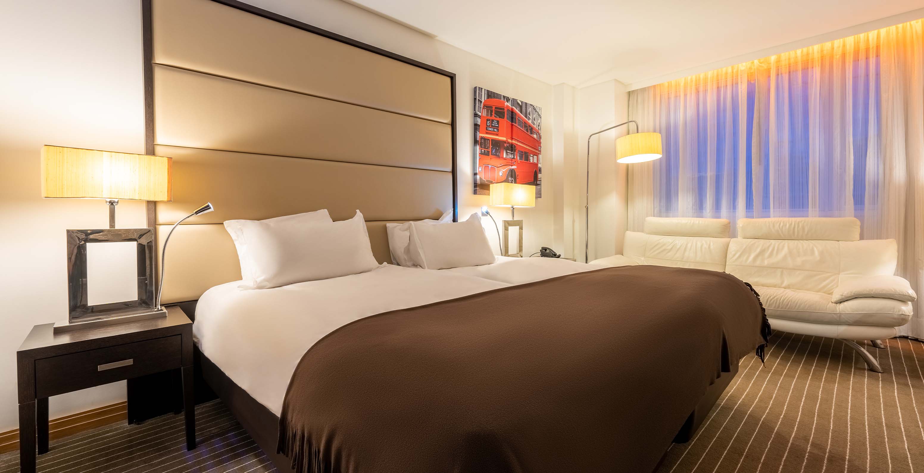 The Deluxe King at Pestana Chelsea Bridge has two joined beds, a bedside table, sofa, lamps, and windows with curtains