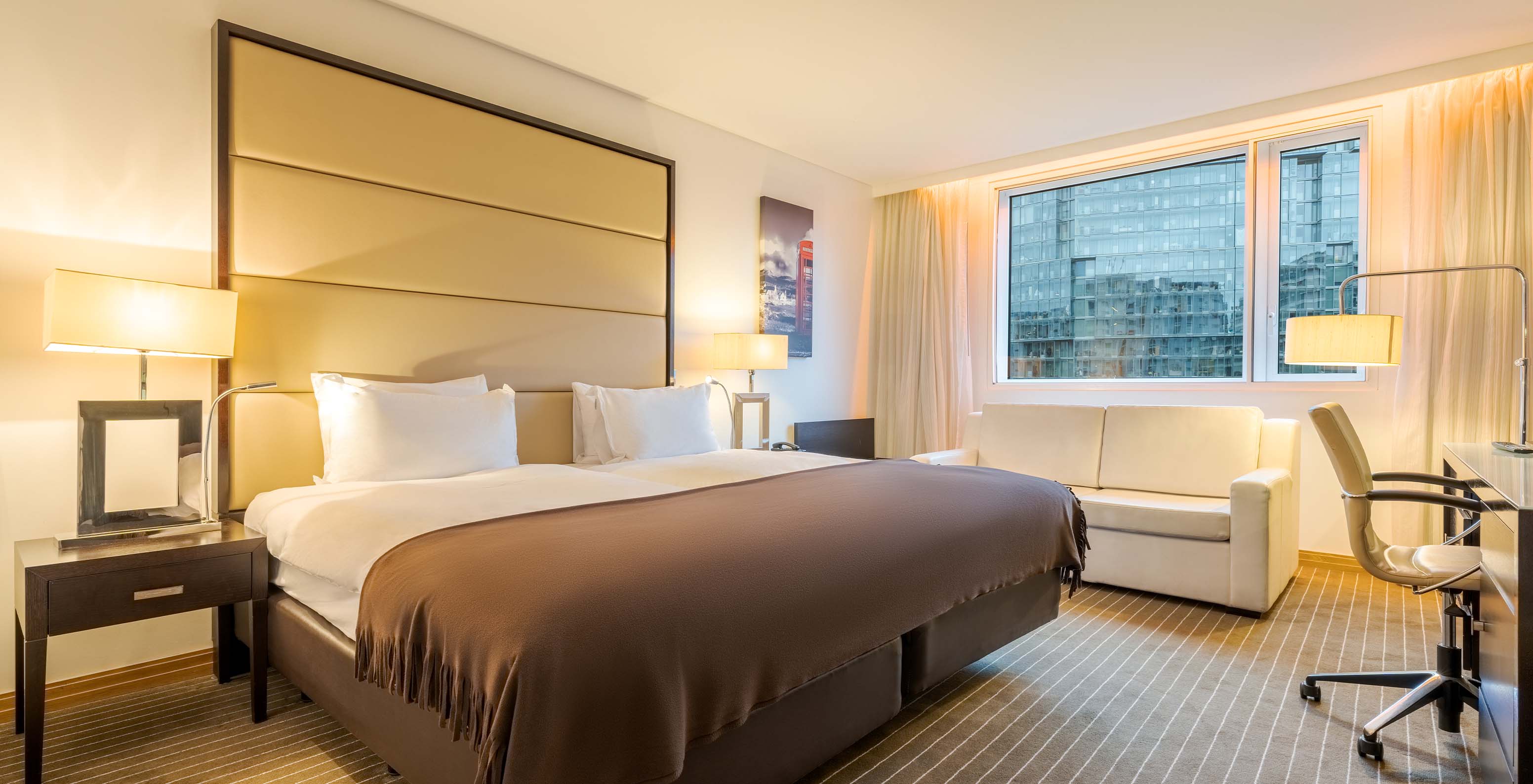 The Twin Deluxe at Pestana Chelsea Bridge has two joined beds, a bedside table, sofa, lamps, and windows with curtains