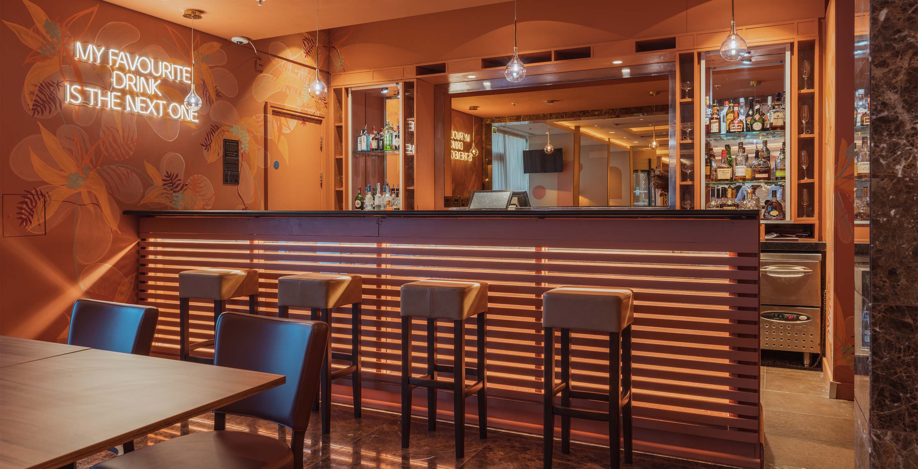 The Bridge bar at the 4-Star Hotel in London has a wooden counter with high stools and a neon phrase on the wall