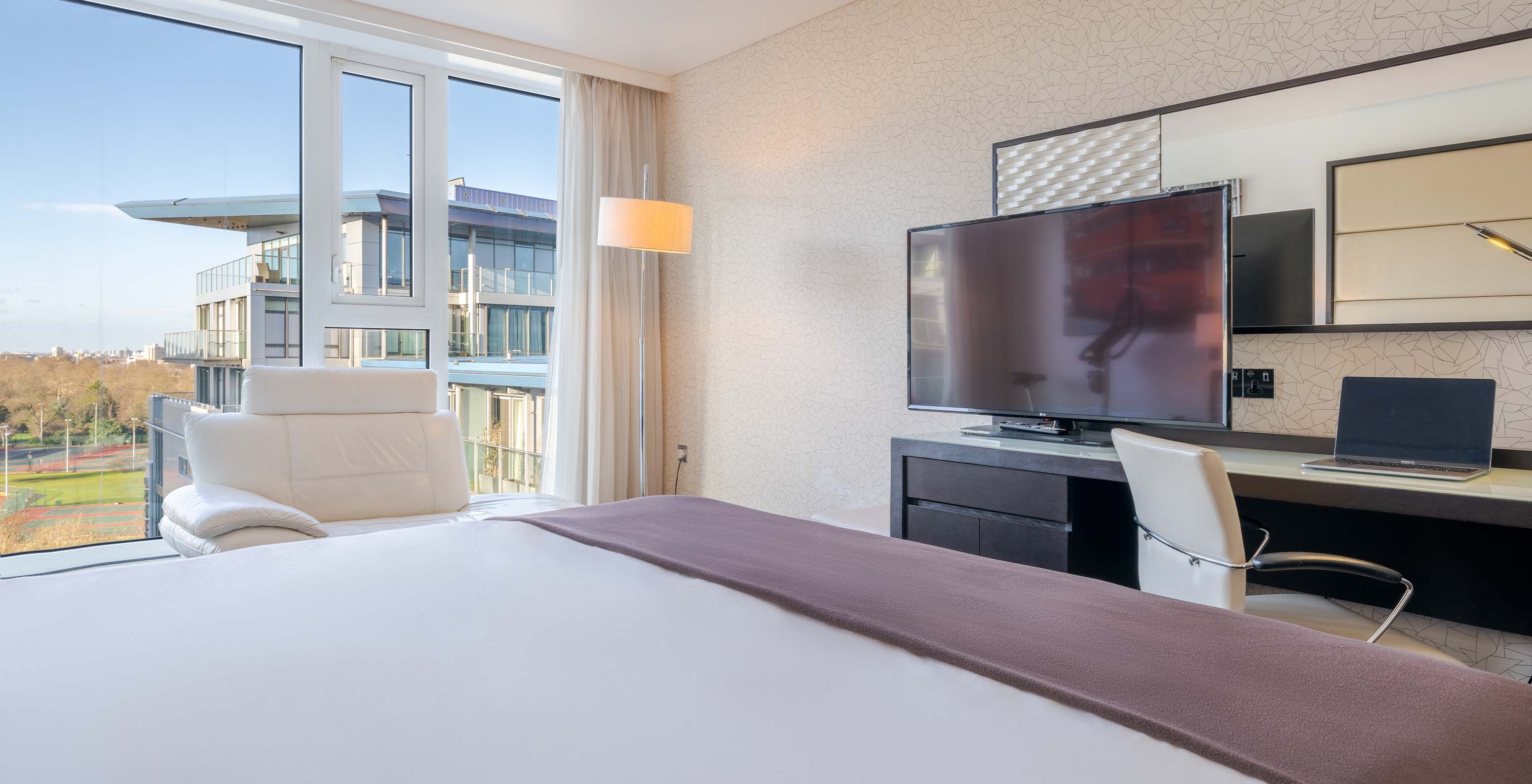 The Deluxe Premium Twin at Pestana Chelsea Bridge has a double bed, a desk with a TV, a sofa, and city windows