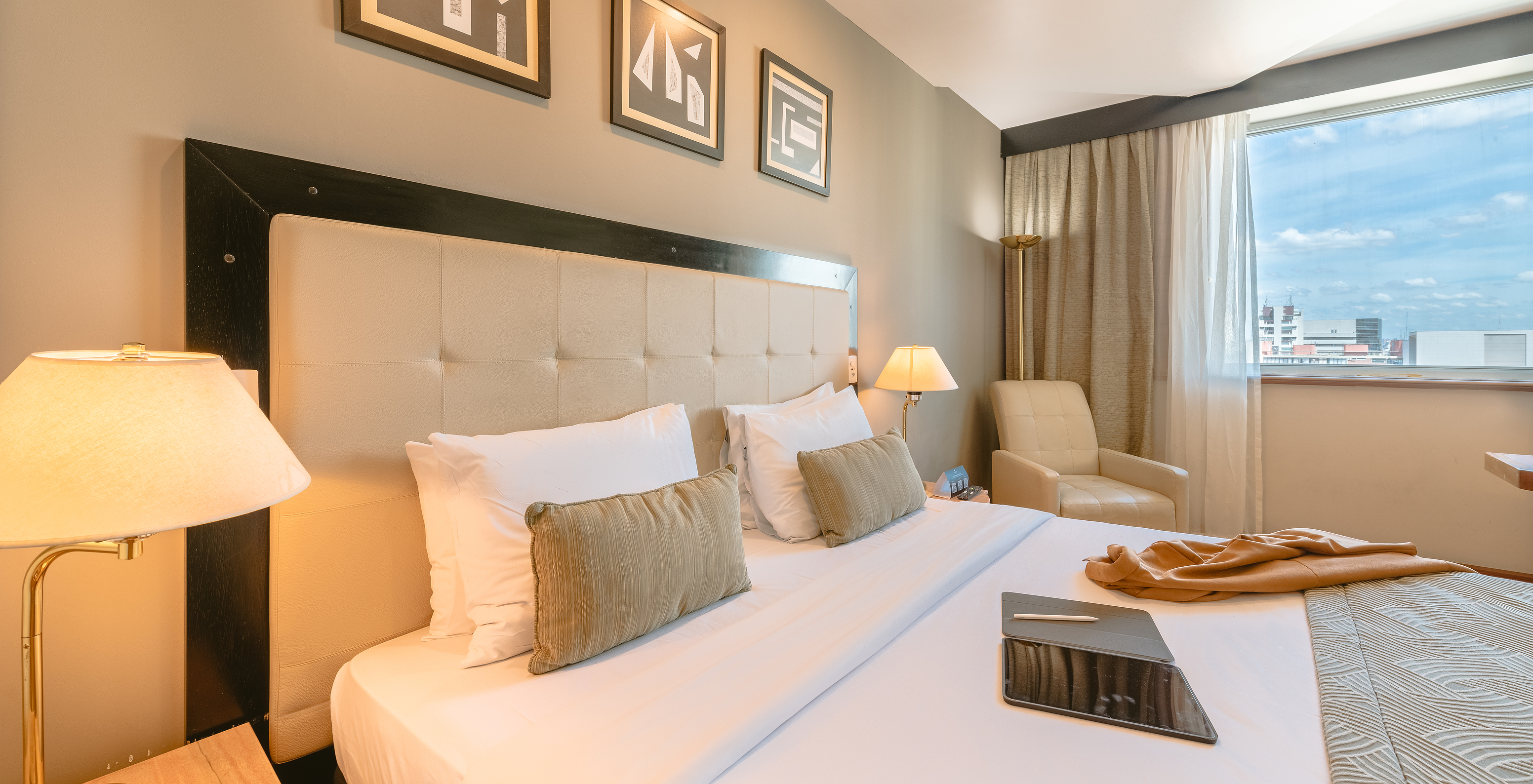 The Standard Room of Pestana São Paulo has a double bed with a beige headboard and a window with city view