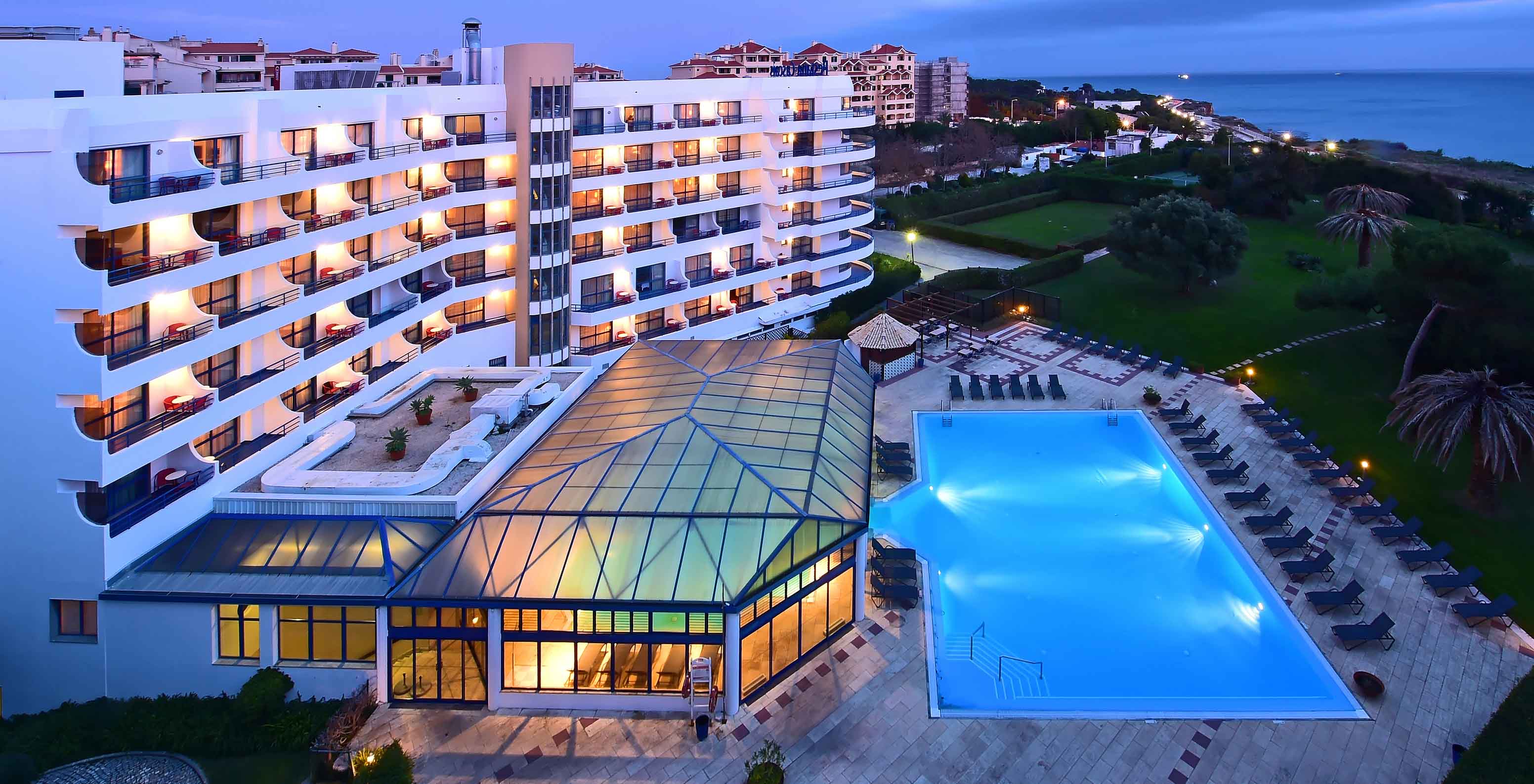 Night view of the hotel, with the room balconies, the garden with the pool, and the sea in the background.