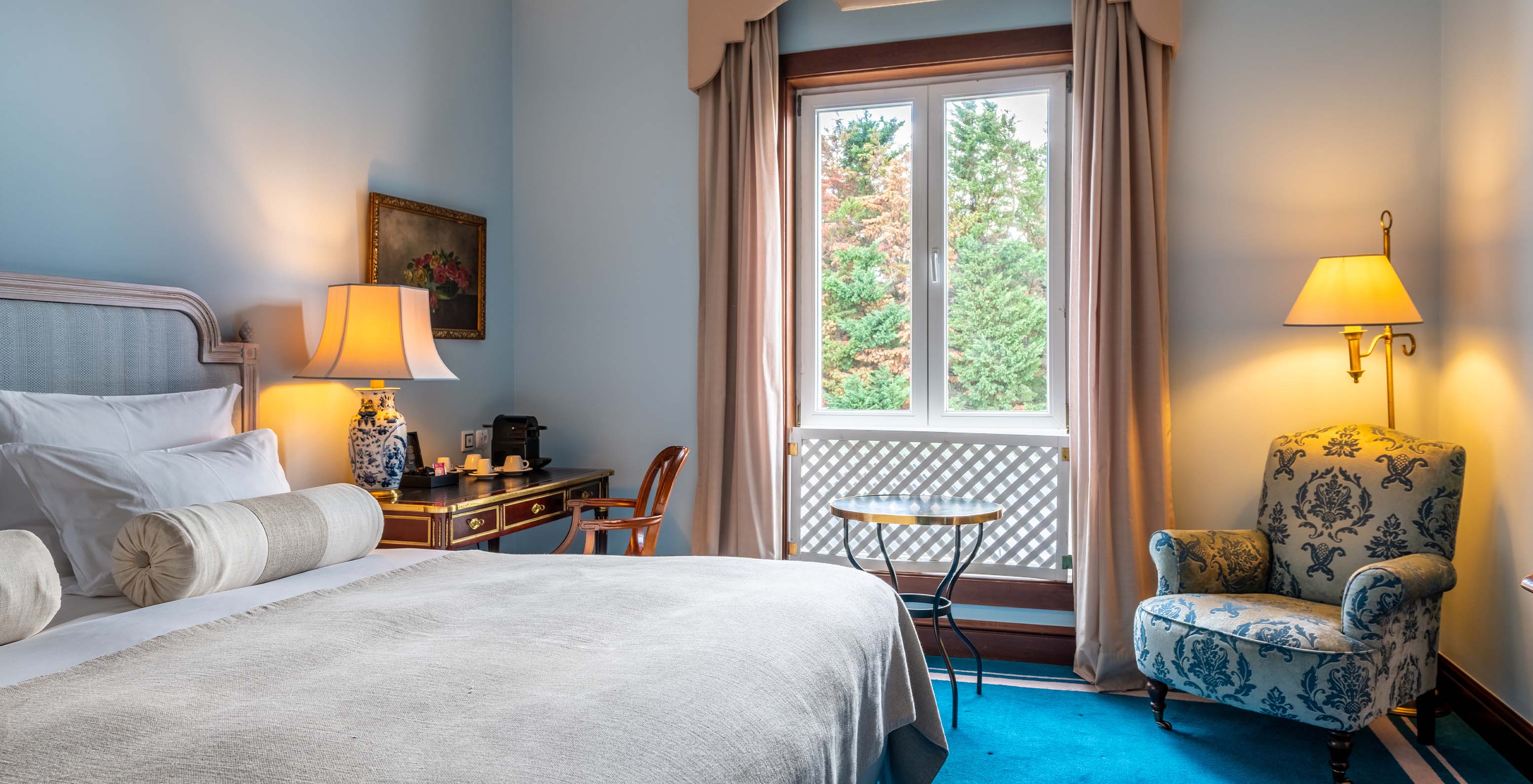 The Deluxe room at Pestana Palace Lisbon includes a double bed, a desk, a window with a decorative grille, and a blue carpet