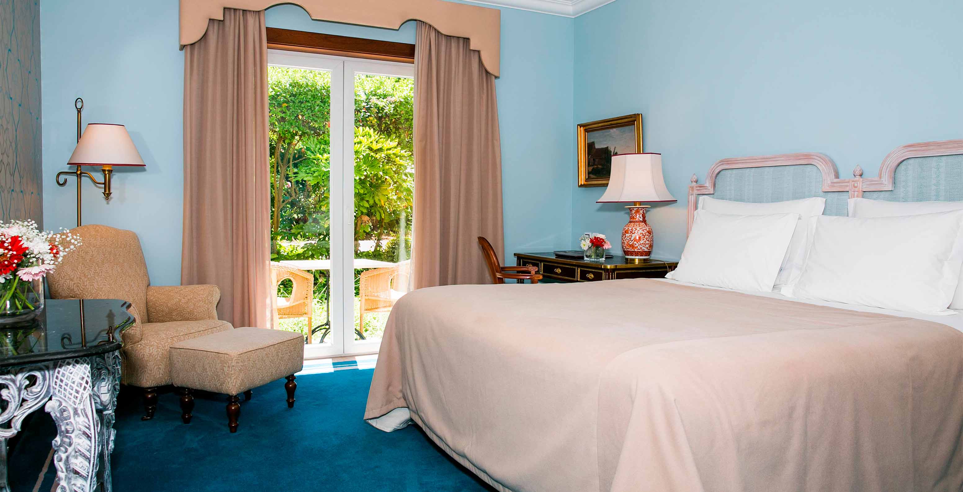 The Suite at Pestana Palace Lisbon has blue walls, a double bed, and a window with access to the garden with a table