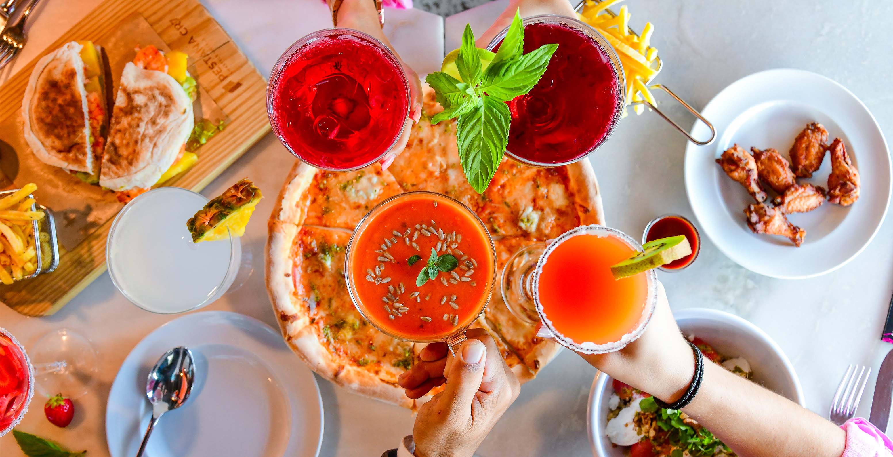 Friends toasting with colorful cocktails in shades of orange, with a pizza in the center and several side dishes