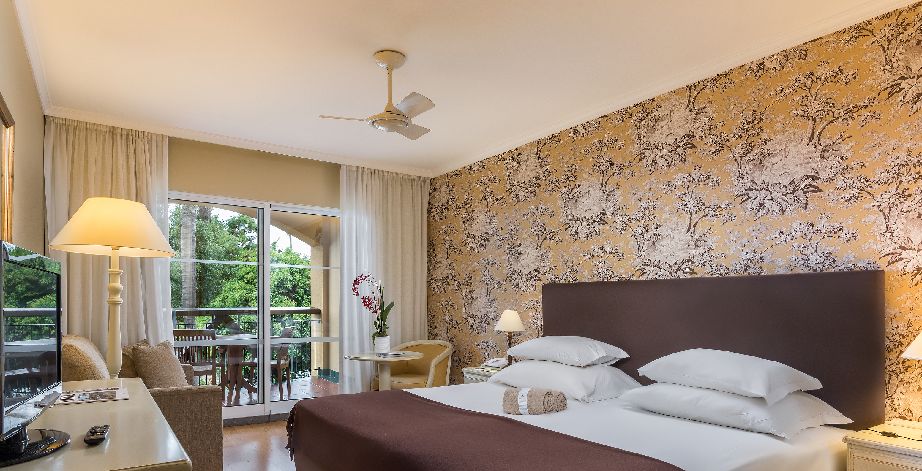 The Studio Classic at Pestana Village has a double bed with headboard and brown bedspread, floral wallpaper and balcony