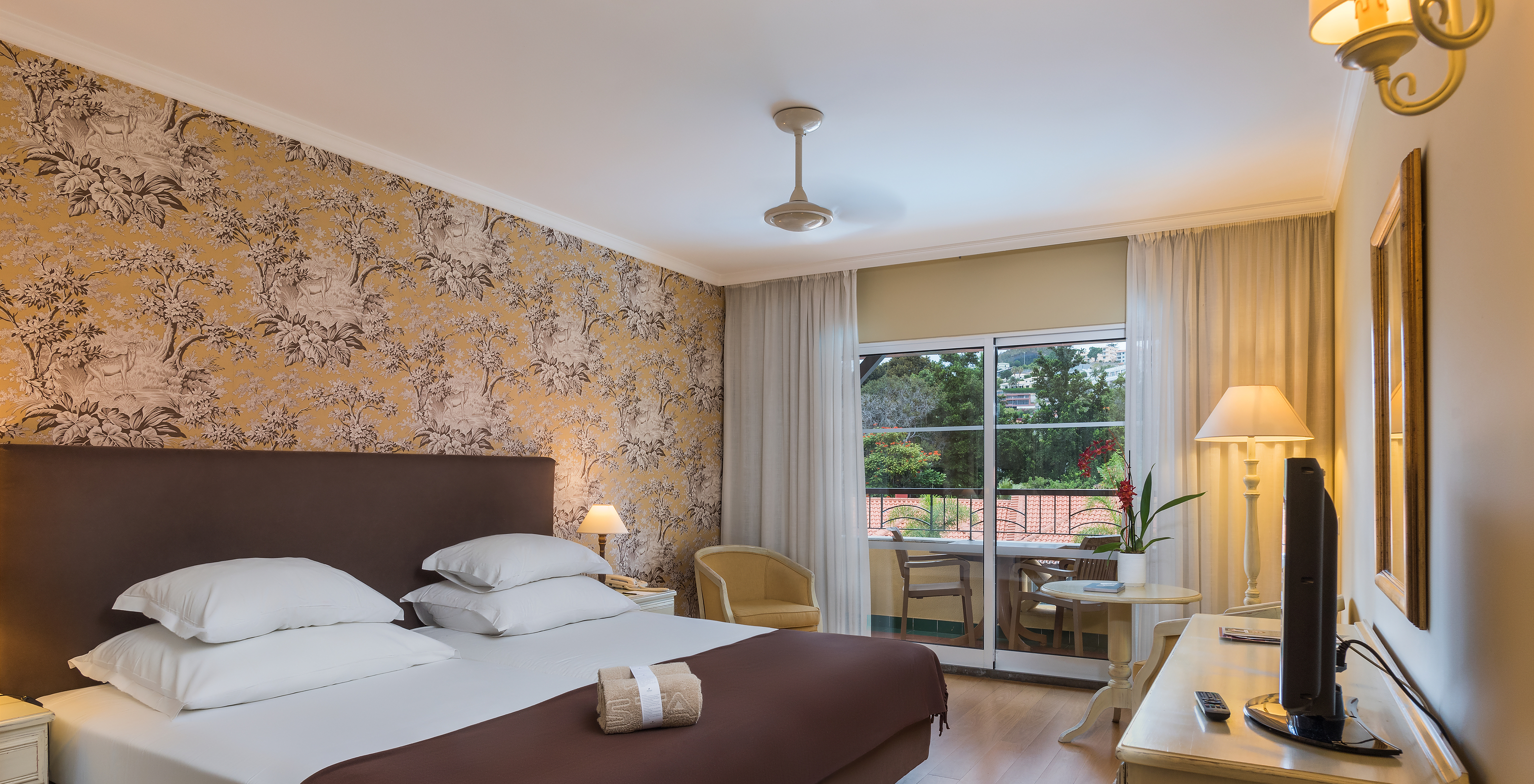 The Studio Superior at Pestana Village has a double bed with headboard and brown bedspread, floral wallpaper and balcony