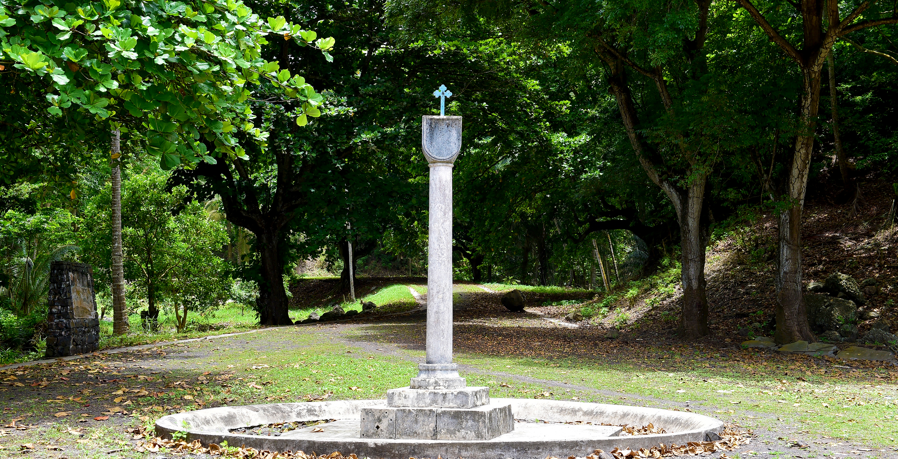 Pathway surrounded by lush trees with a stone monument in the center, topped by a cross