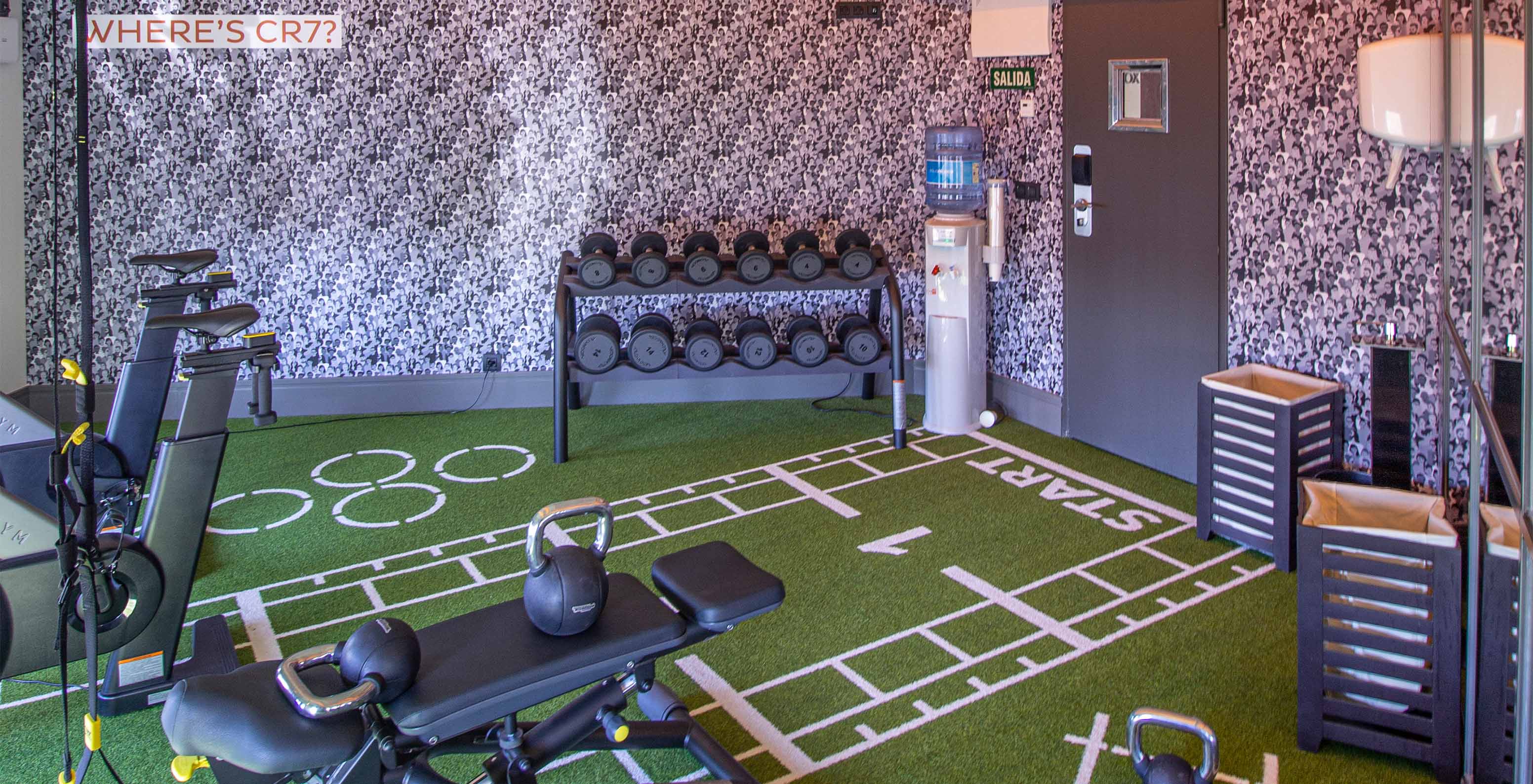 Gym with various weights, dumbbells, bicycles, benches, TRX, and towel baskets, with green grass-like flooring