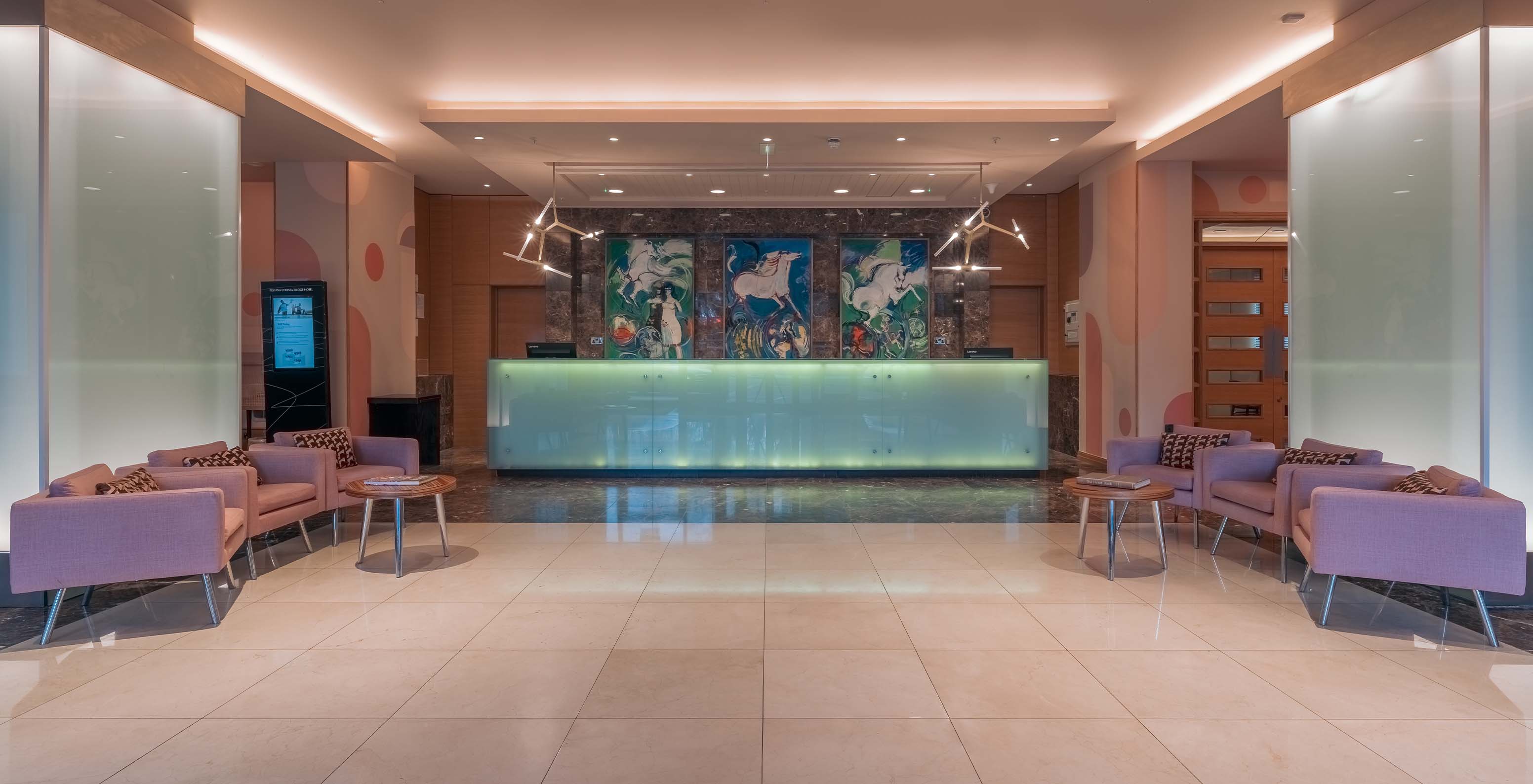 Lobby reception area of the hotel, with purple sofas, side tables, a lit counter, colorful paintings on the wall, and lamps