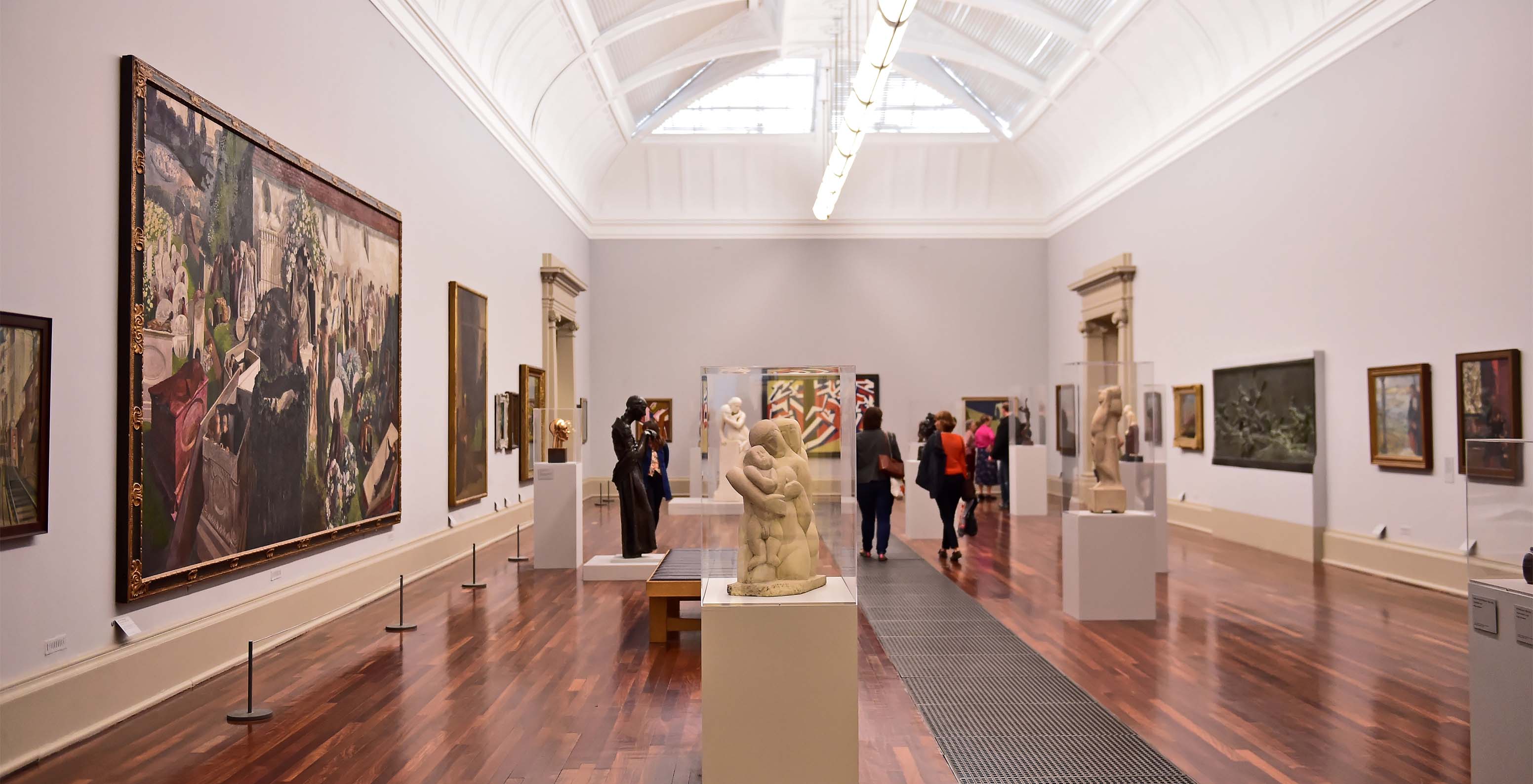 Museum room with paintings, sculptures, people walking, wooden floor, and white walls