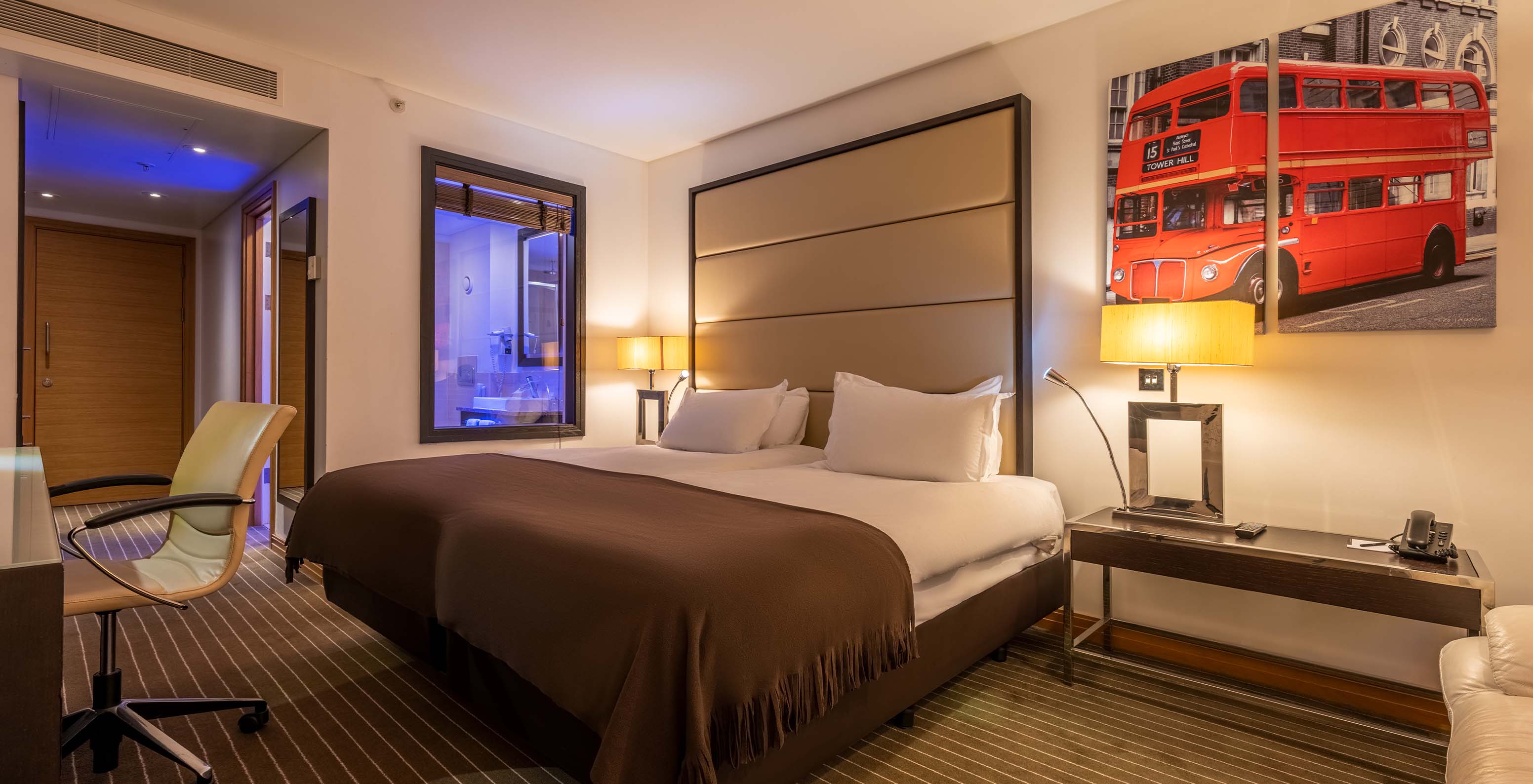 The Deluxe King at Pestana Chelsea Bridge has two beds with London pictures on the wall, a bedside table, sofa, and chair