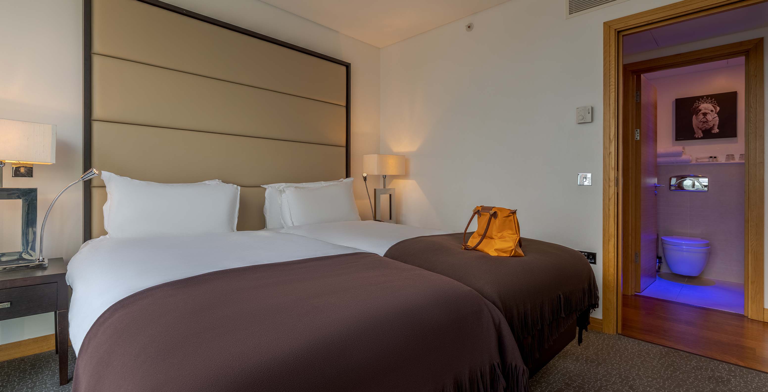 The Deluxe Premium Twin at Pestana Chelsea Bridge has two joined beds, bedside tables with lamps, and a bathroom