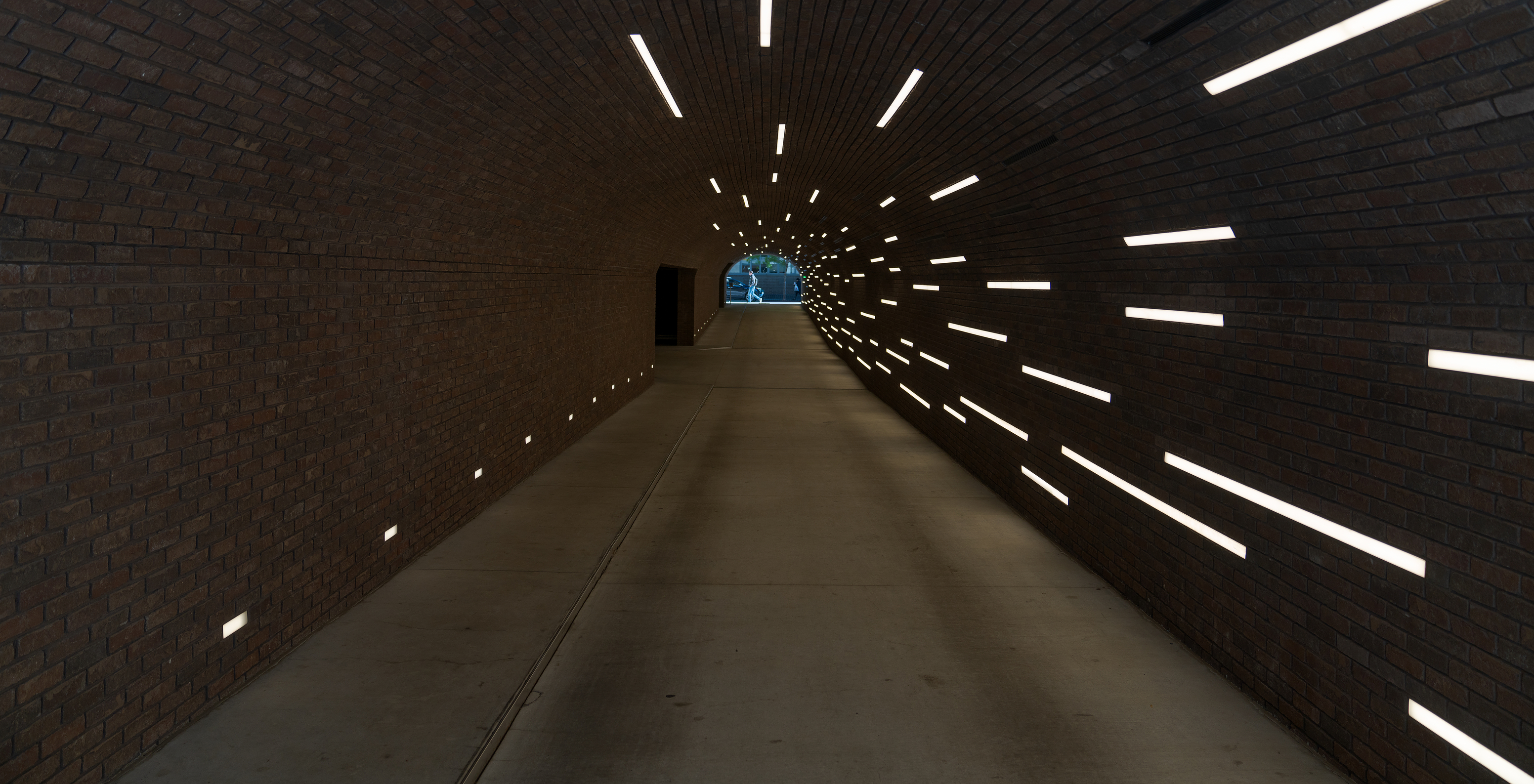 View of an interior tunnel with concrete floor and brick walls illuminated by fluorescent light lines