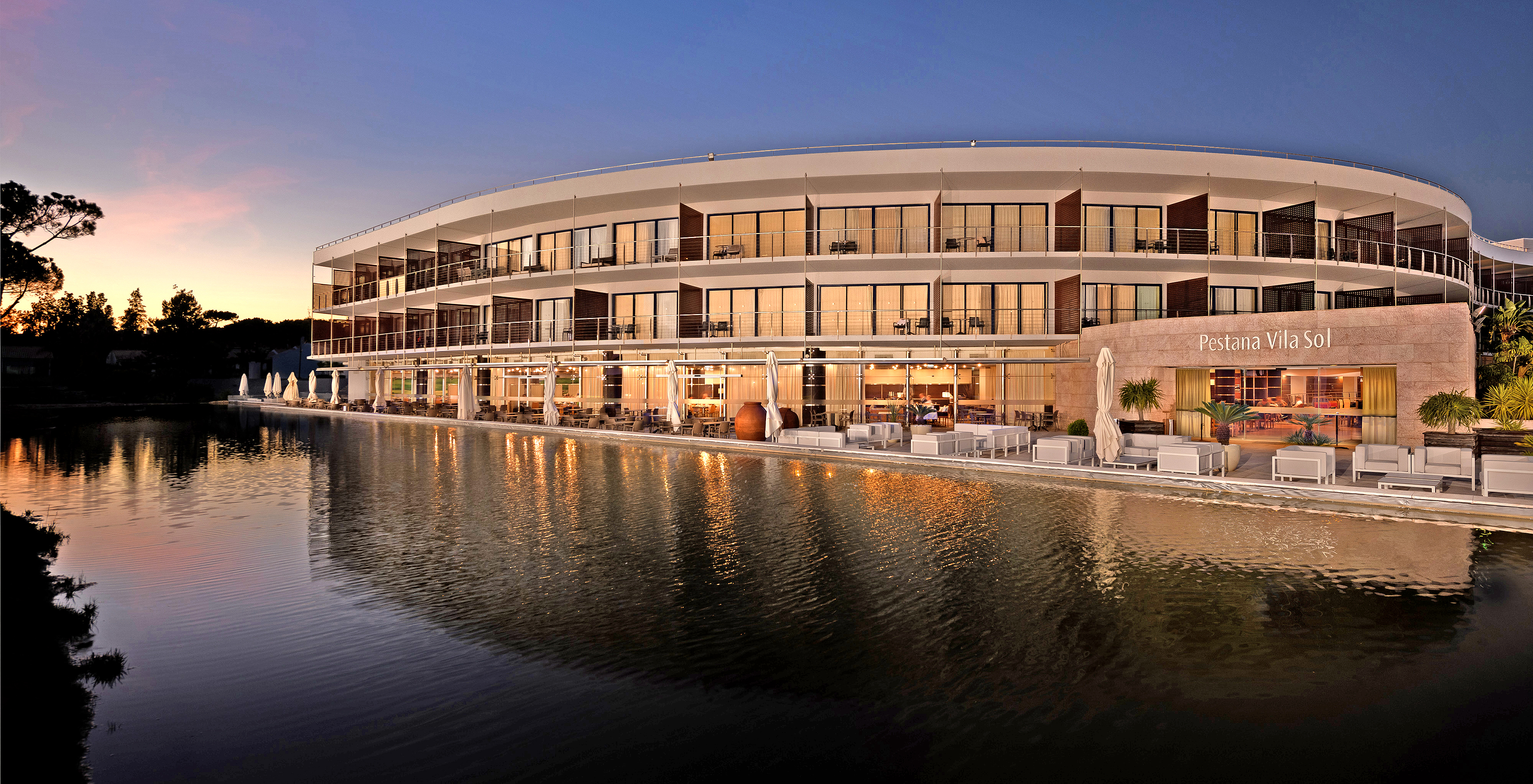 Vista del edificio del Pestana Vila Sol - Vilamoura al atardecer, con un lago enfrente y varias mesas y sofás en la terraza