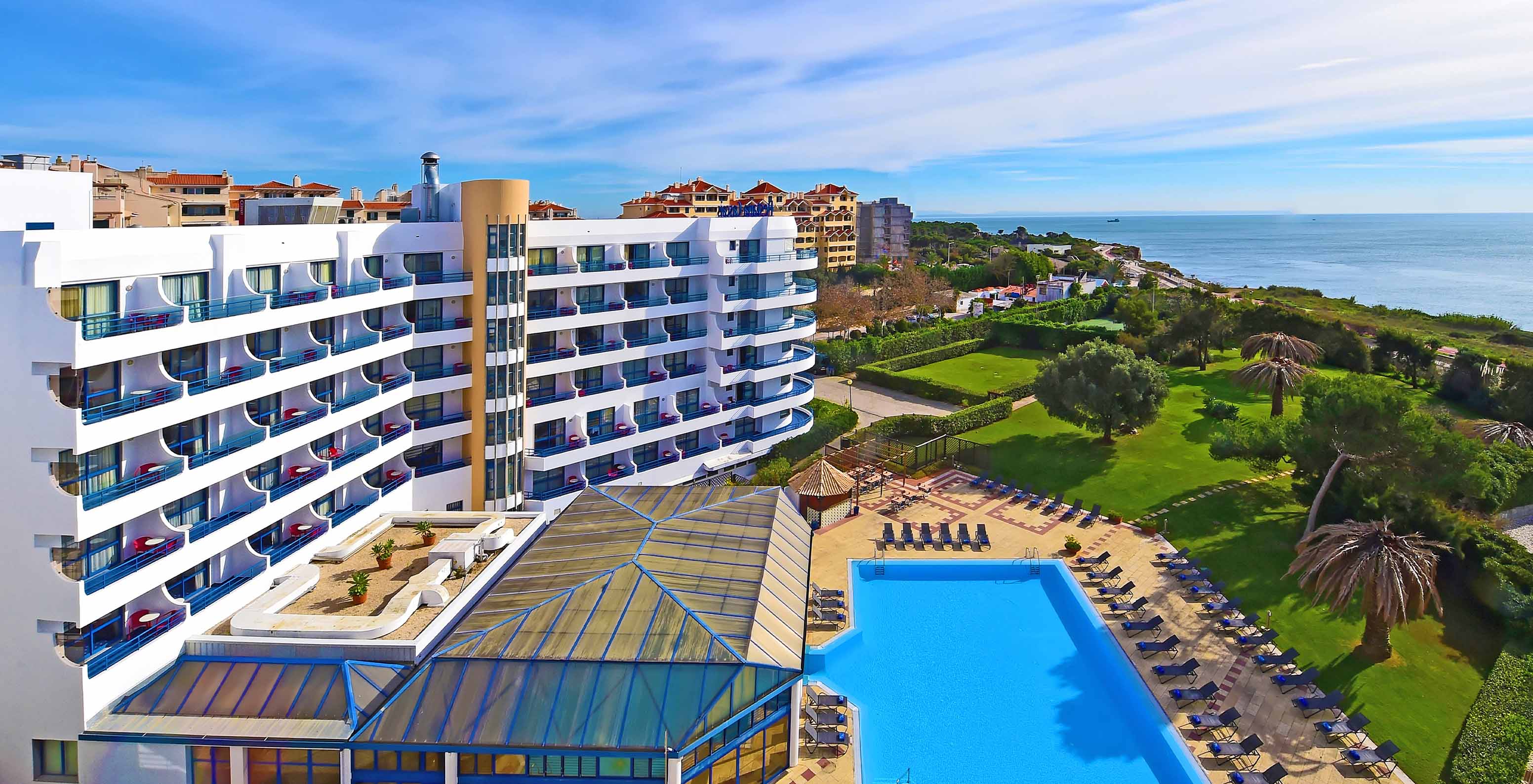 Vista exterior del Pestana Cascais desde la piscina, fachada del hotel y campos de césped con palmeras