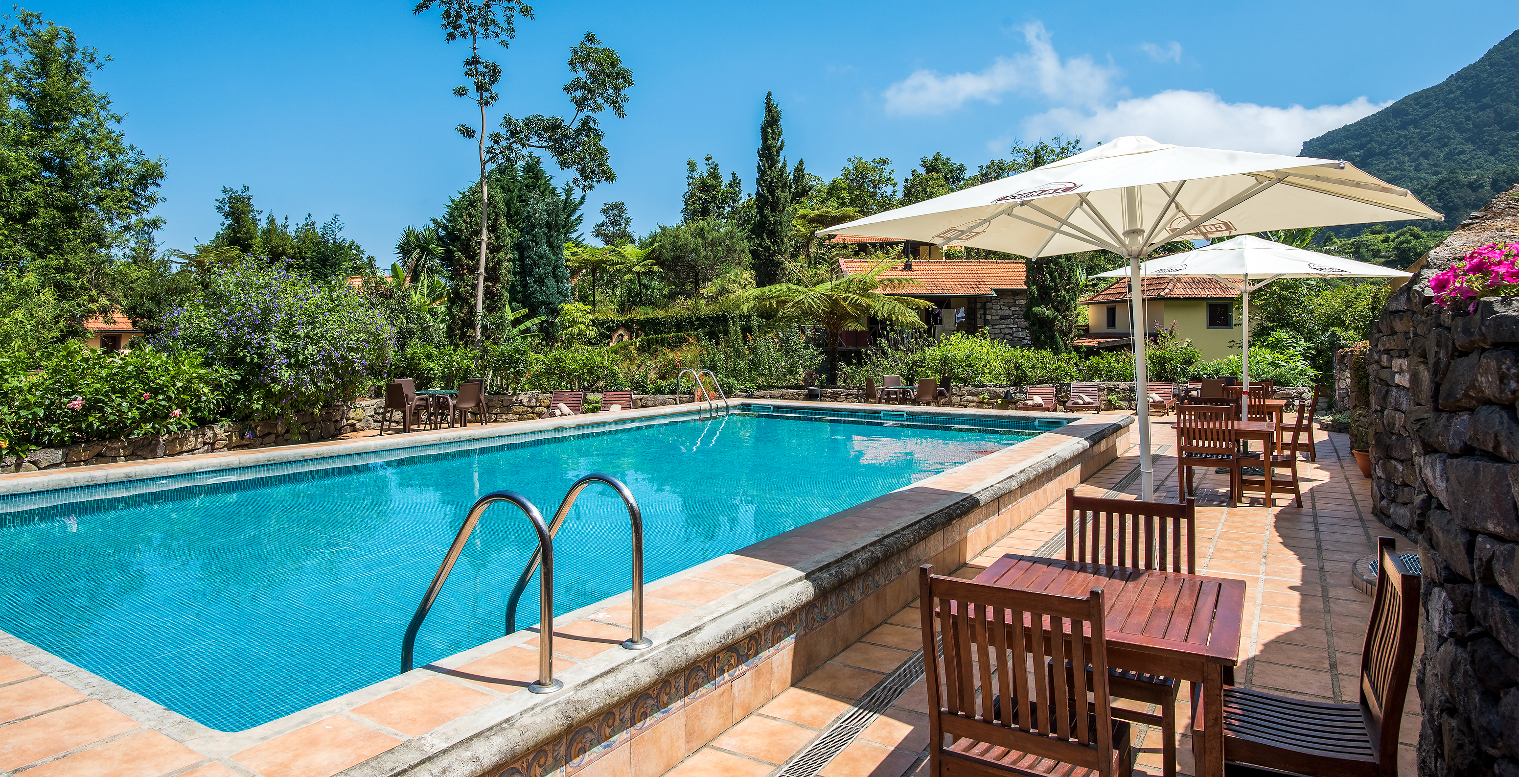 Piscina exterior con mesas, sillas, tumbonas y sombrillas, con las villas y vegetación al fondo